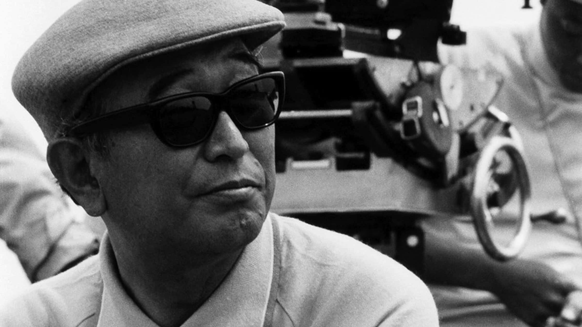 Akira Kurosawa 桌布 壁紙 — 一個戴帽和太陽眼鏡的男人的黑白肖像，背景有一部菲林相機。