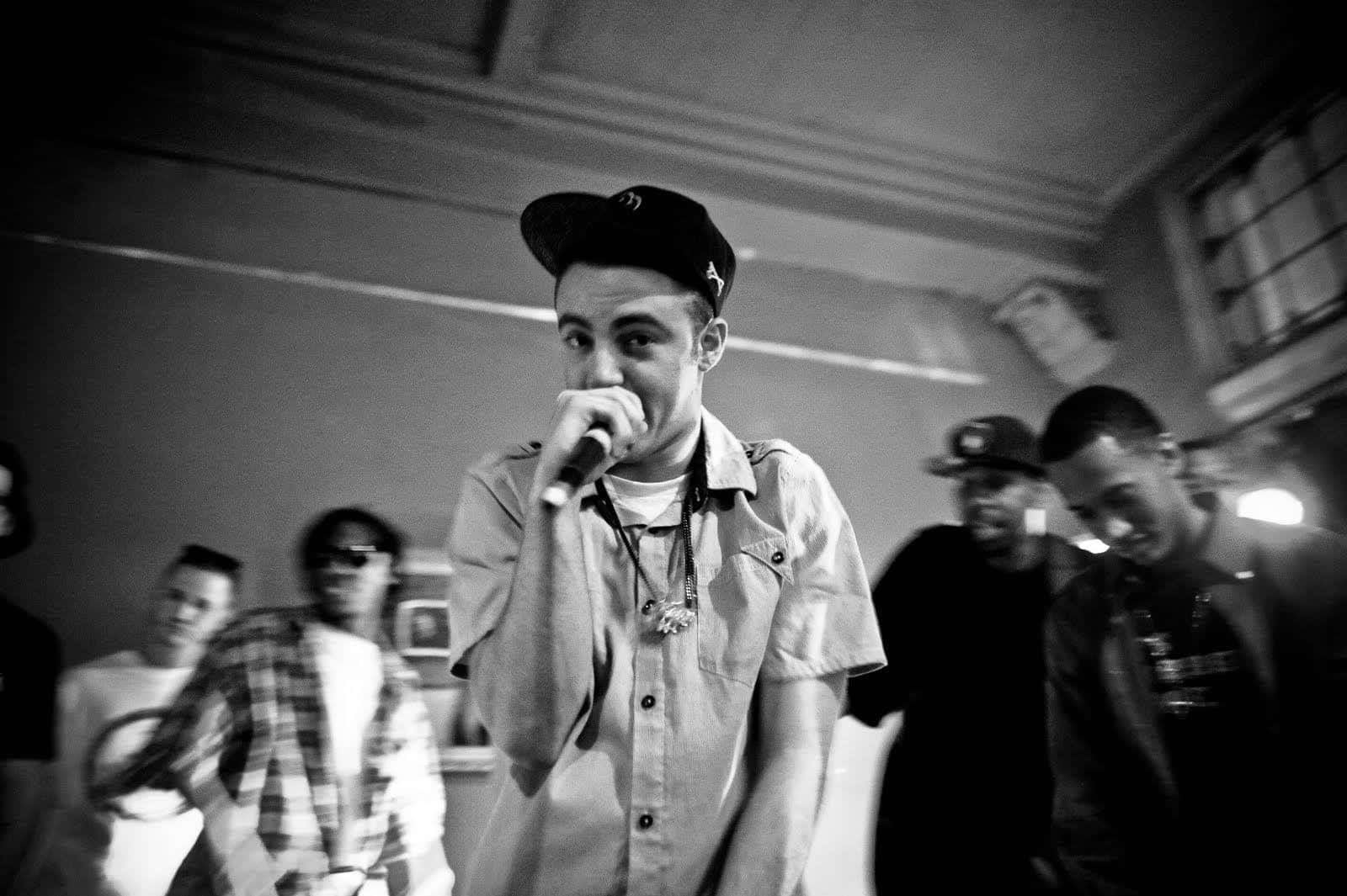 Mac Miller 桌布 壁紙 — 一個戴帽嘅年輕男子熱情咁對住咪高峰rap，模糊嘅朋友喺昏暗嘅房間睇住。