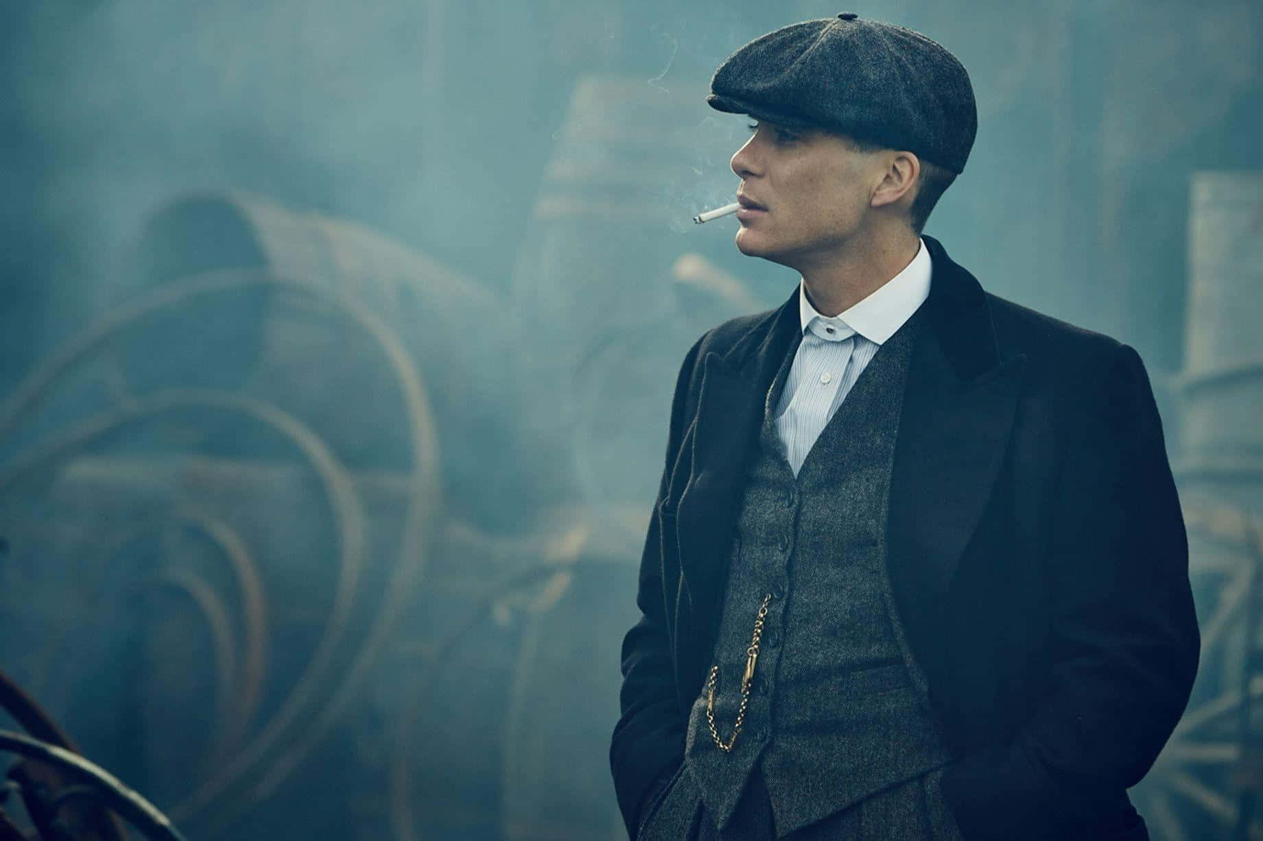 Peaky Blinders 桌布 壁紙 — 一個戴平頂帽、身穿復古西裝的男人，在煙霧瀰漫的工業背景下抽煙。