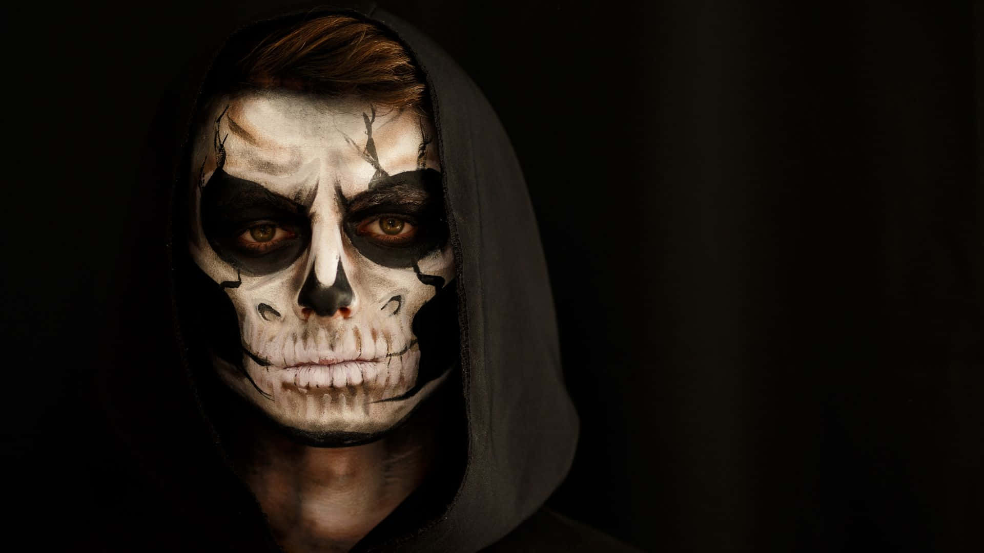 Horror Costumes 桌布 壁紙 — 一個戴深色連帽衫的人，在戲劇性燈光下畫著帶有裂骨效果的骷髏妝。