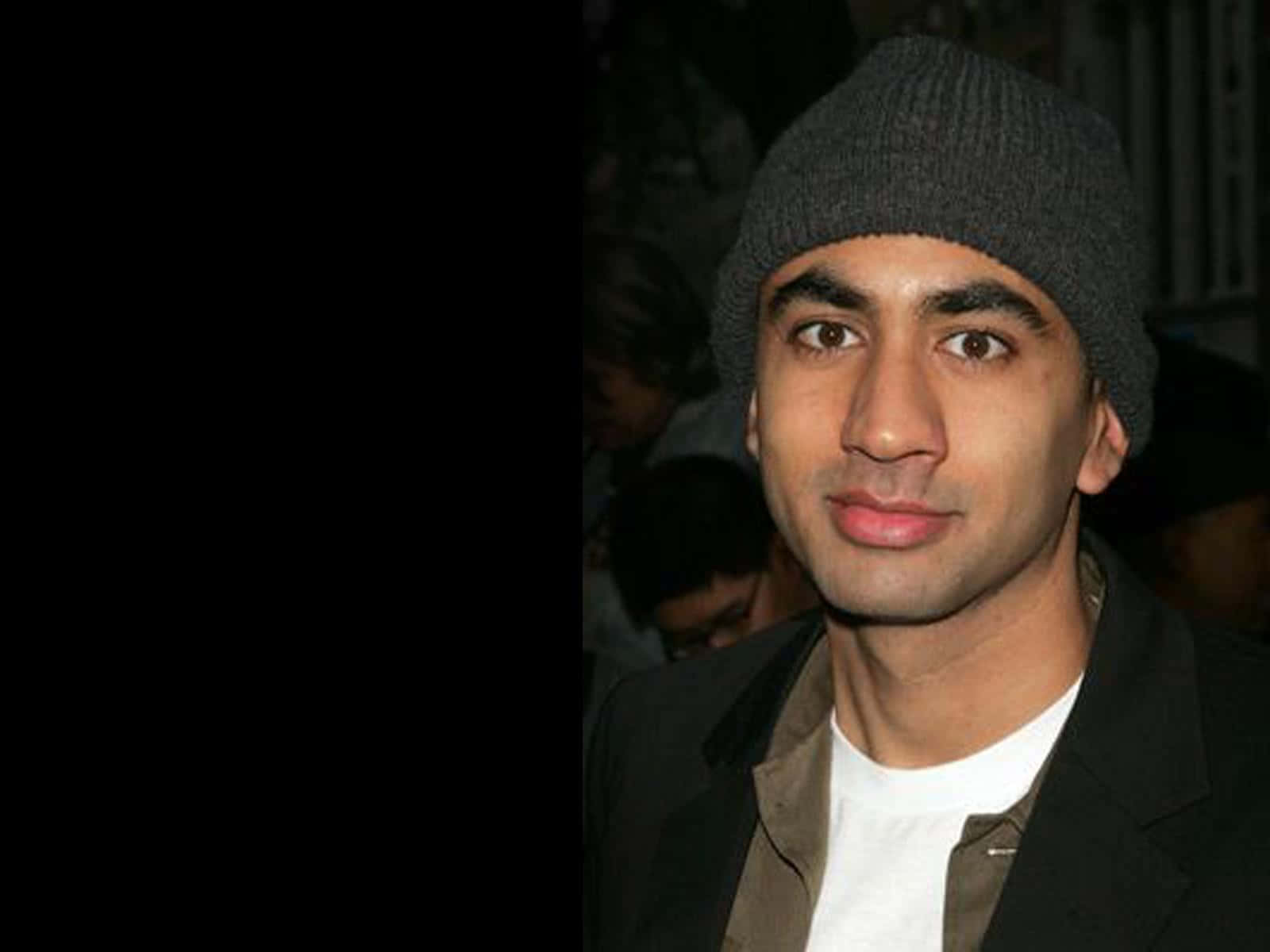 Kal Penn 桌布 壁紙 — 一個戴灰色冷帽、穿深色外套的男人，在昏暗的人群背景下輕輕微笑。