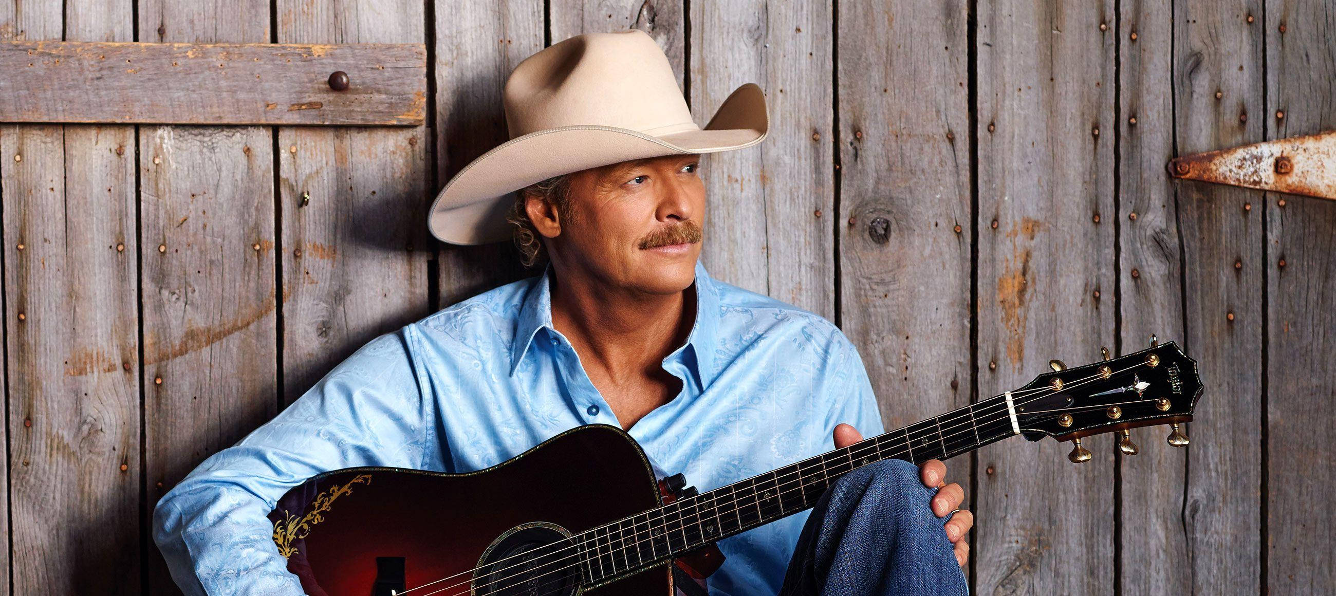 Alan Jackson 桌布 壁紙 — 一個戴牛仔帽、穿藍色恤衫的男人靠著風化木頭，手持木結他。