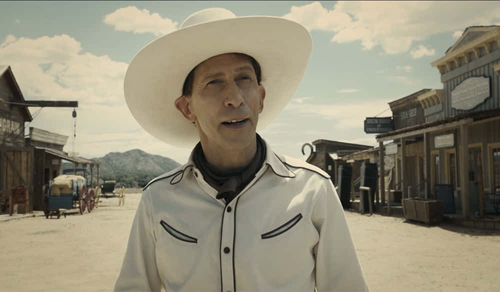 Tim Blake Nelson 桌布 壁紙 — 一個戴白色牛仔帽、穿白色襯衫的男人站在塵土飛揚的西部小鎮街道上，背景有木造建築和山脈，天空局部多雲。