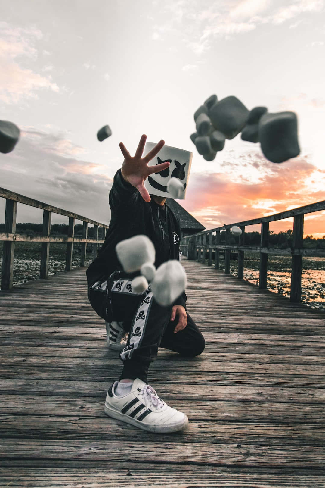 Marshmallow 桌布 壁紙 — 一個戴白色面具、穿黑色衣服的人蹲在木碼頭上，在日落時將石頭拋向空中。