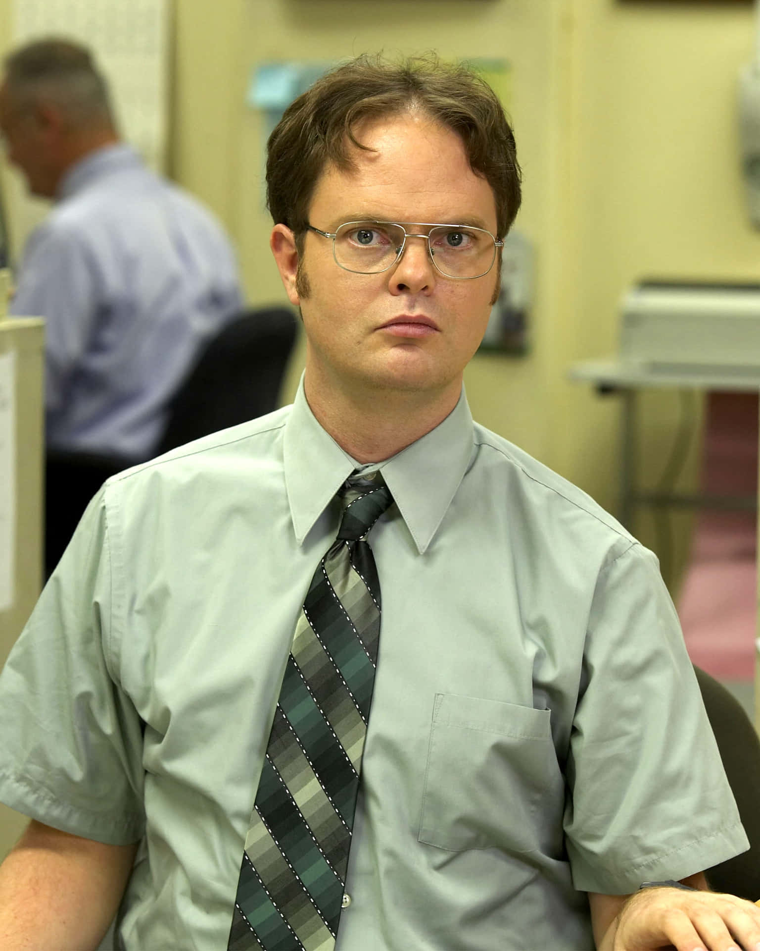 rainn wilson 桌布 壁紙 — 一個戴眼鏡、打條紋領帶的男人坐在辦公室隔間裡，直視鏡頭，表情嚴肅。