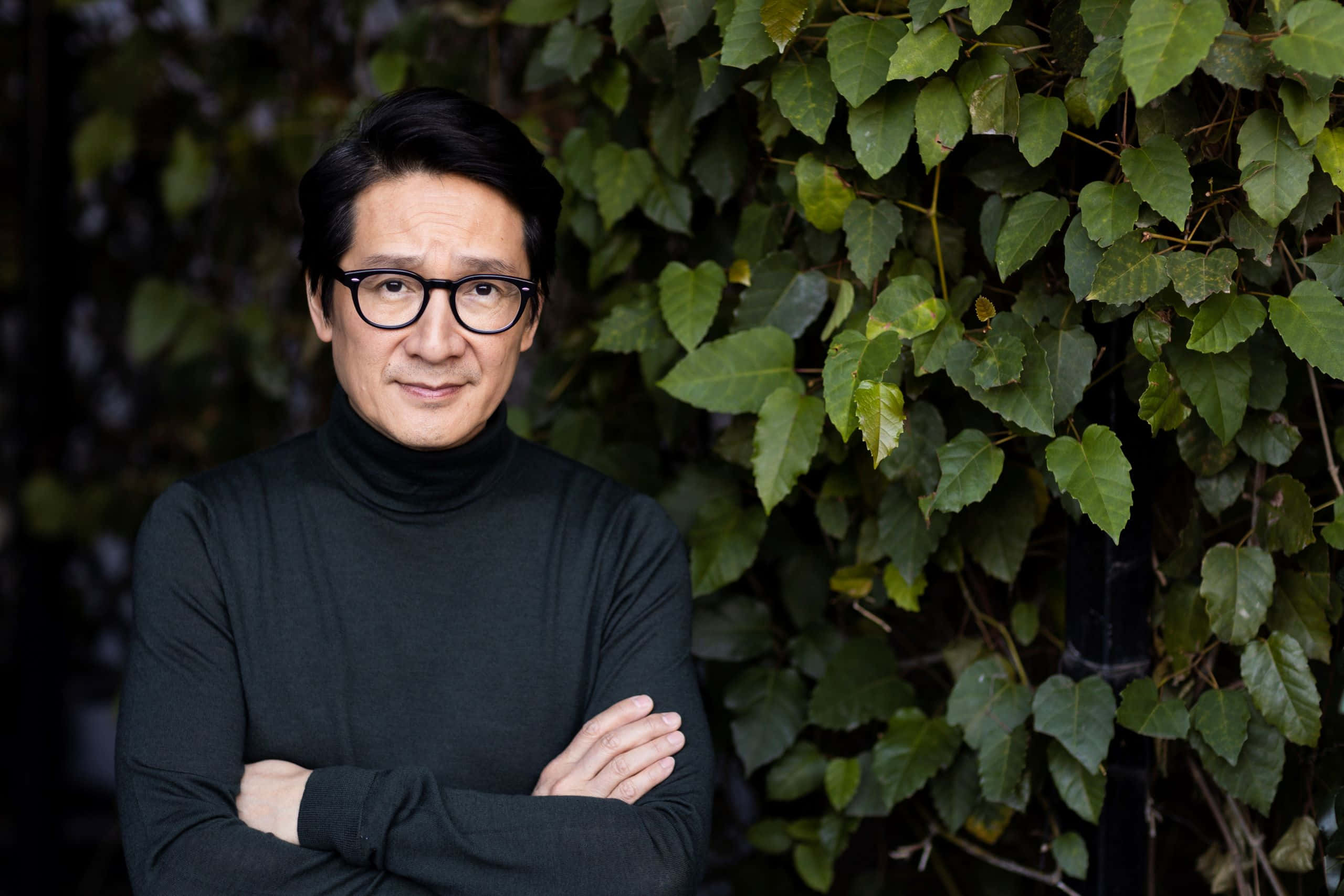 Ke Huy Quan 桌布 壁紙 — 一個戴眼鏡、穿黑色高領毛衣的男人自信地交叉雙臂，靠在一面茂密的綠色常春藤牆上。