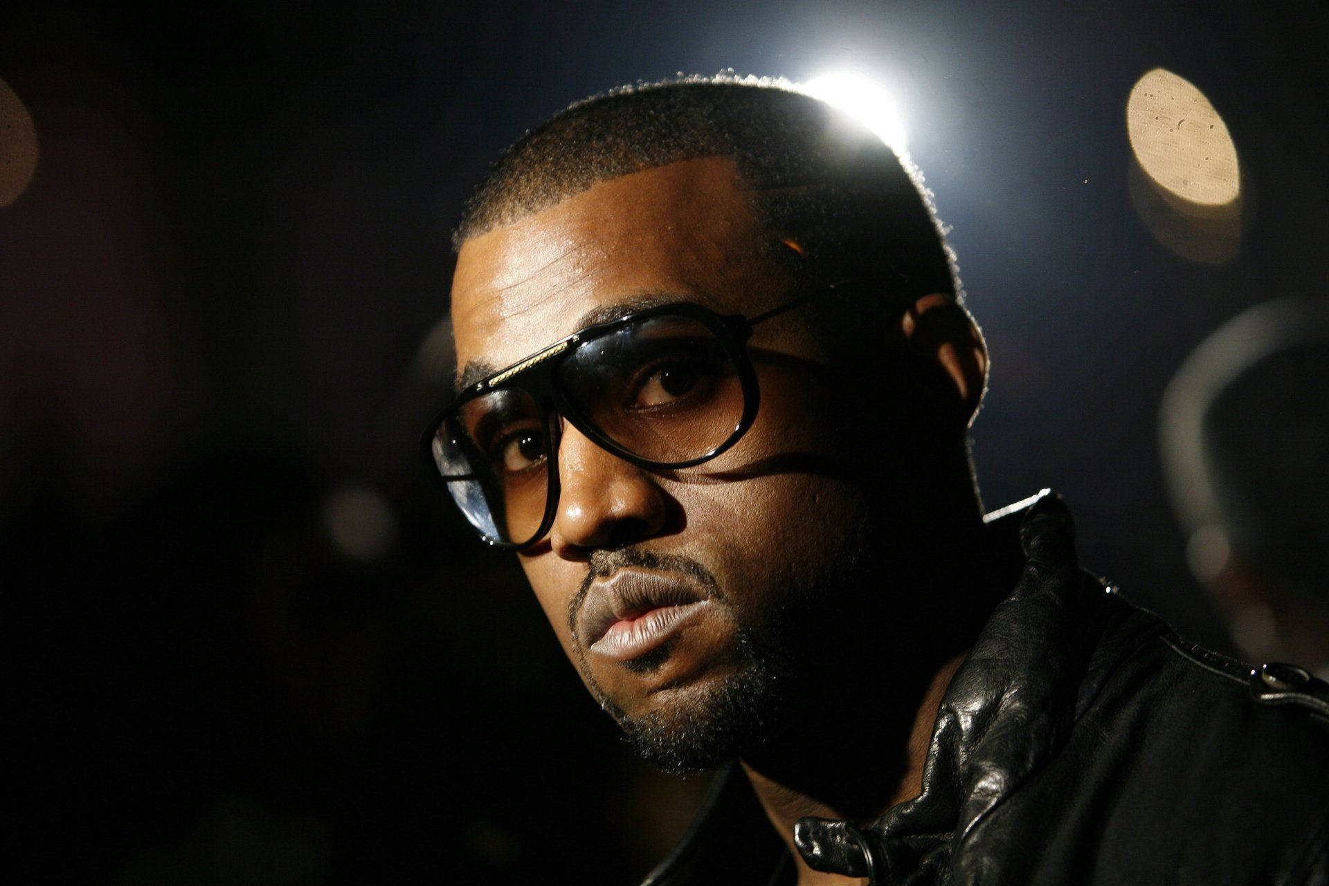 Kanye West Brown Tint Glasses 壁紙 — 一個戴眼鏡和皮褸的男人在戲劇性舞台燈光下，背景有散景效果，他凝視著。