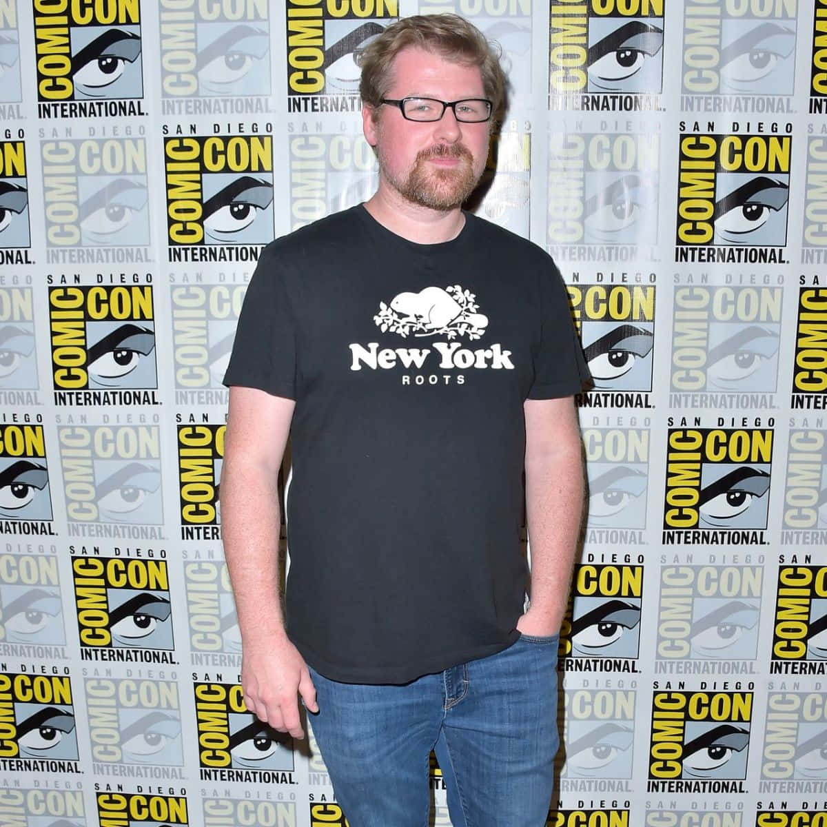 Justin Roiland 桌布 壁紙 — 一個戴眼鏡和鬍鬚的男人自信地站在重複的Comic-Con International背景前，穿著黑色'New York Roots' T恤和牛仔褲。