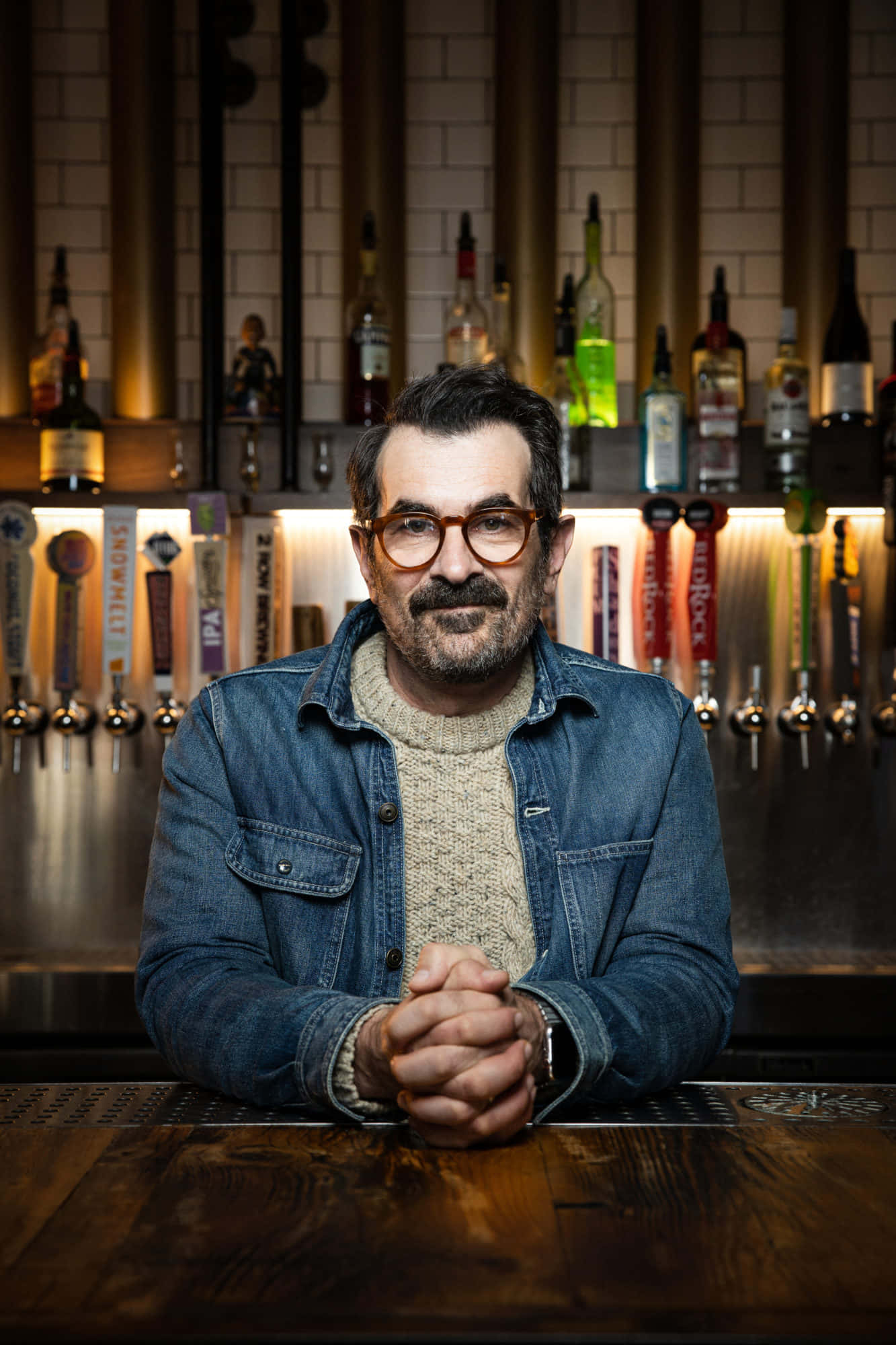 Ty Burrell 桌布 壁紙 — 一個戴眼鏡留鬍鬚的男人坐在木製吧檯前雙手交握，身後有啤酒龍頭和架子上的瓶子。
