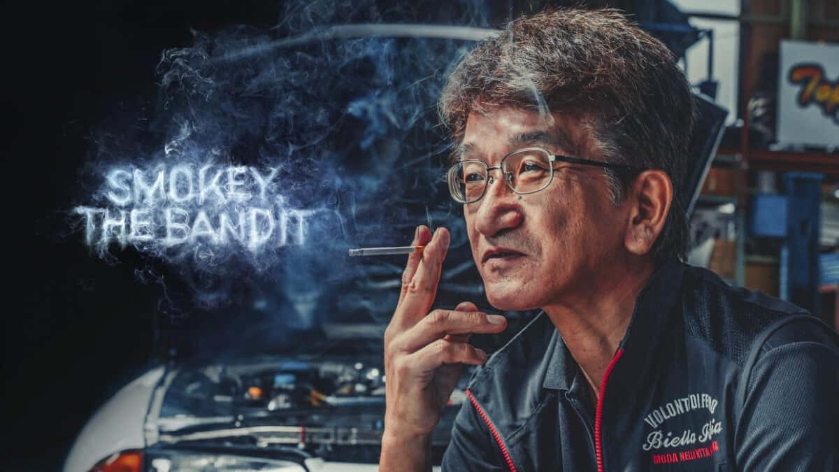 Smokey Nagata The Bandit 壁紙 — 一個戴眼鏡的男人吸煙，煙霧在車庫的汽車上方形成「SMOKEY THE BANDIT」字樣。
