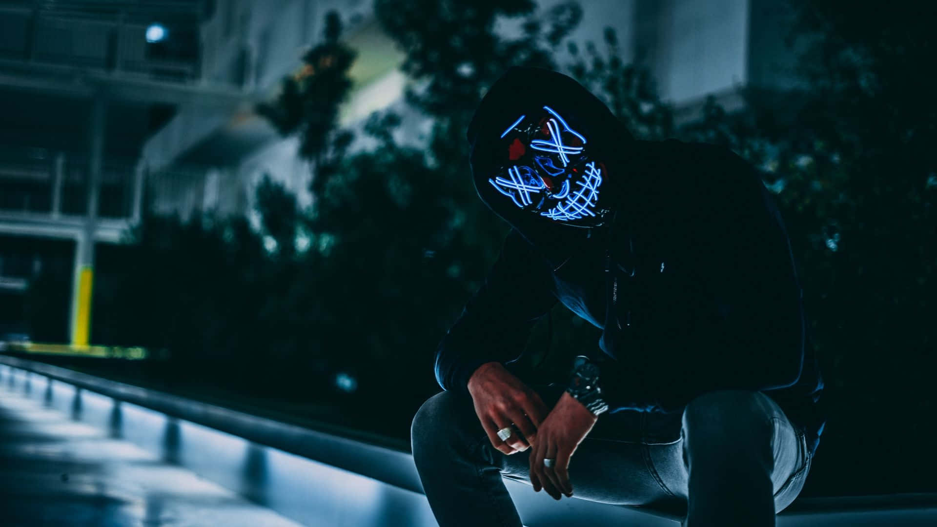 Mask 桌布 壁紙 — 一個戴着發光藍色LED面具的人，在夜晚昏暗的路燈下，獨自蹲在濕漉漉的城市邊緣。