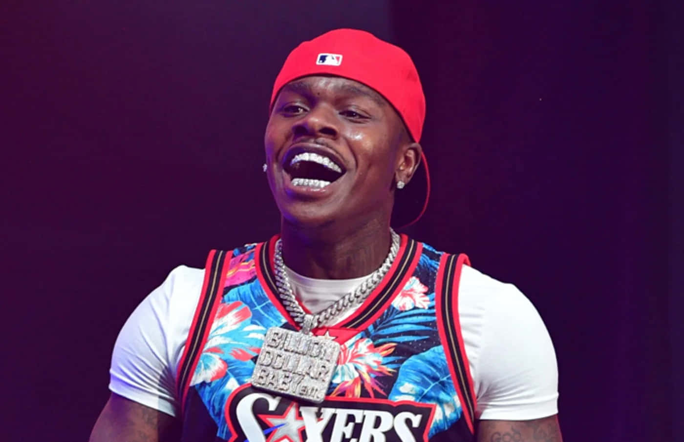 Dababy 桌布 壁紙 — 一個戴紅帽、穿花紋球衣嘅男人喺深紫色背景嘅舞台燈光下開心大笑。