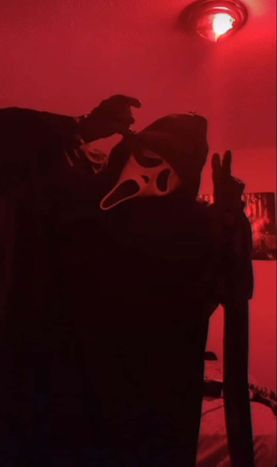 Ghostface Aesthetic 桌布 壁紙 — 一個戴著Ghostface面具的人物站在昏暗的紅色燈光房間中，單手做出和平手勢。