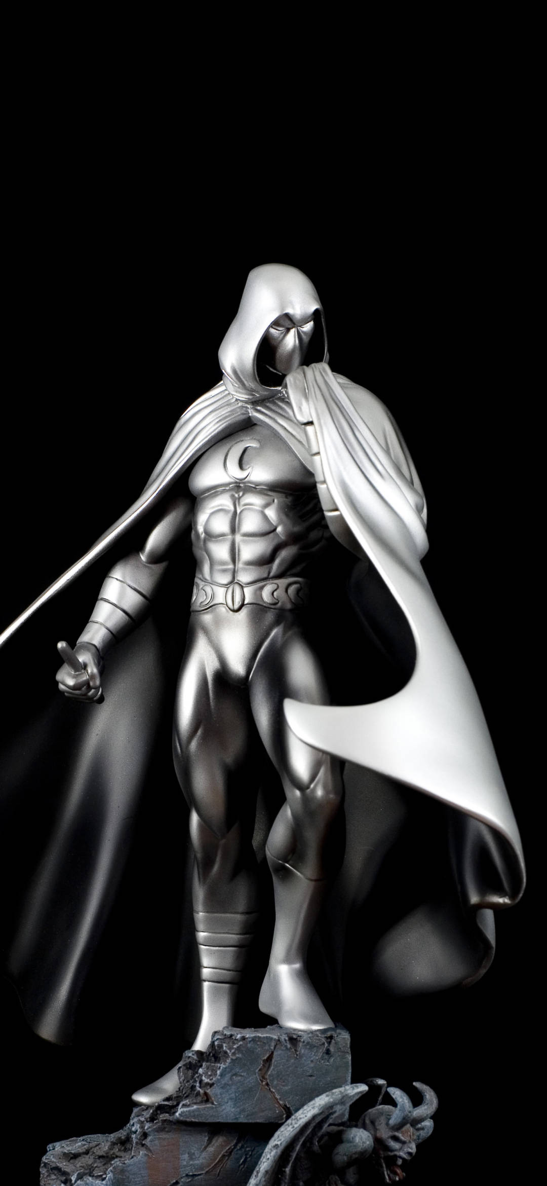 Moon Knight 桌布 壁紙 — 一個戴著兜帽的超級英雄的金屬銀色雕像，披著飄逸的斗篷，站在黑色背景下的岩石底座上。