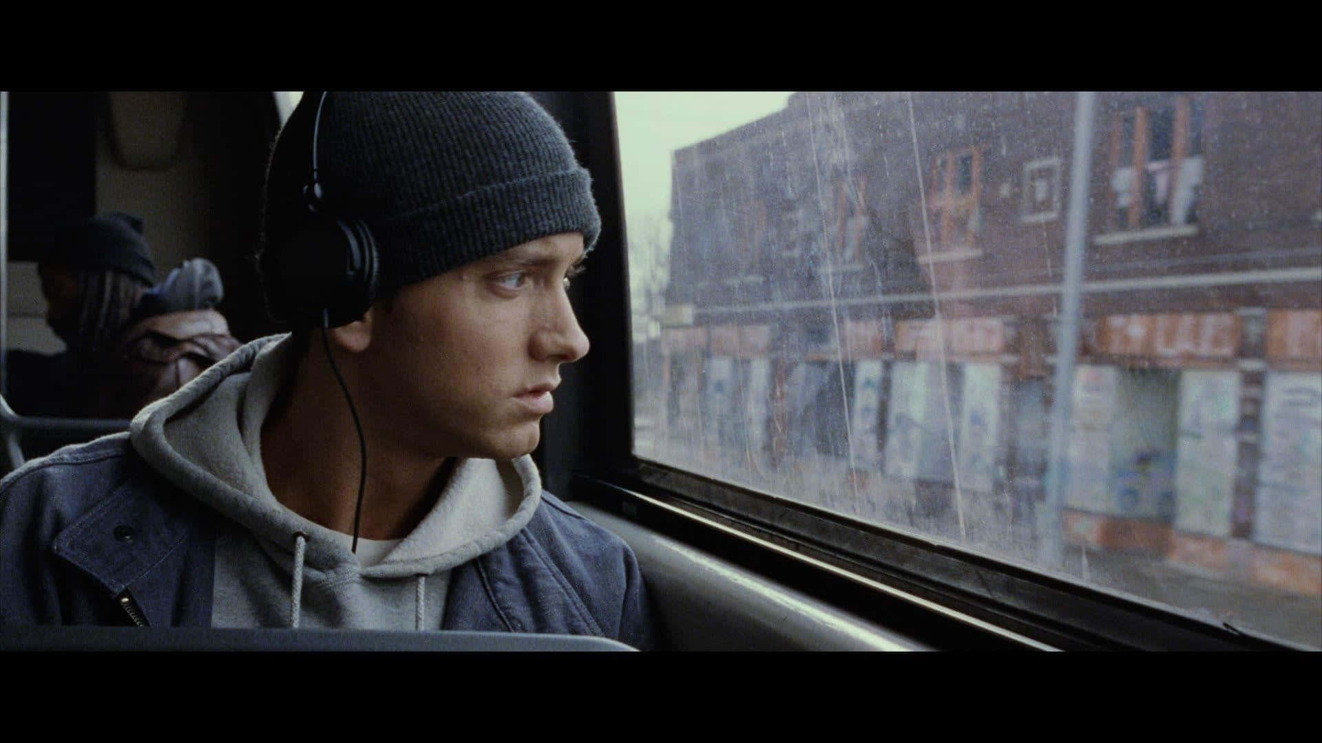 Eminem 桌布 壁紙 — 一個戴著冷帽和耳機的年輕男子，透過佈滿雨痕的巴士車窗凝視著模糊的城市商店門面。