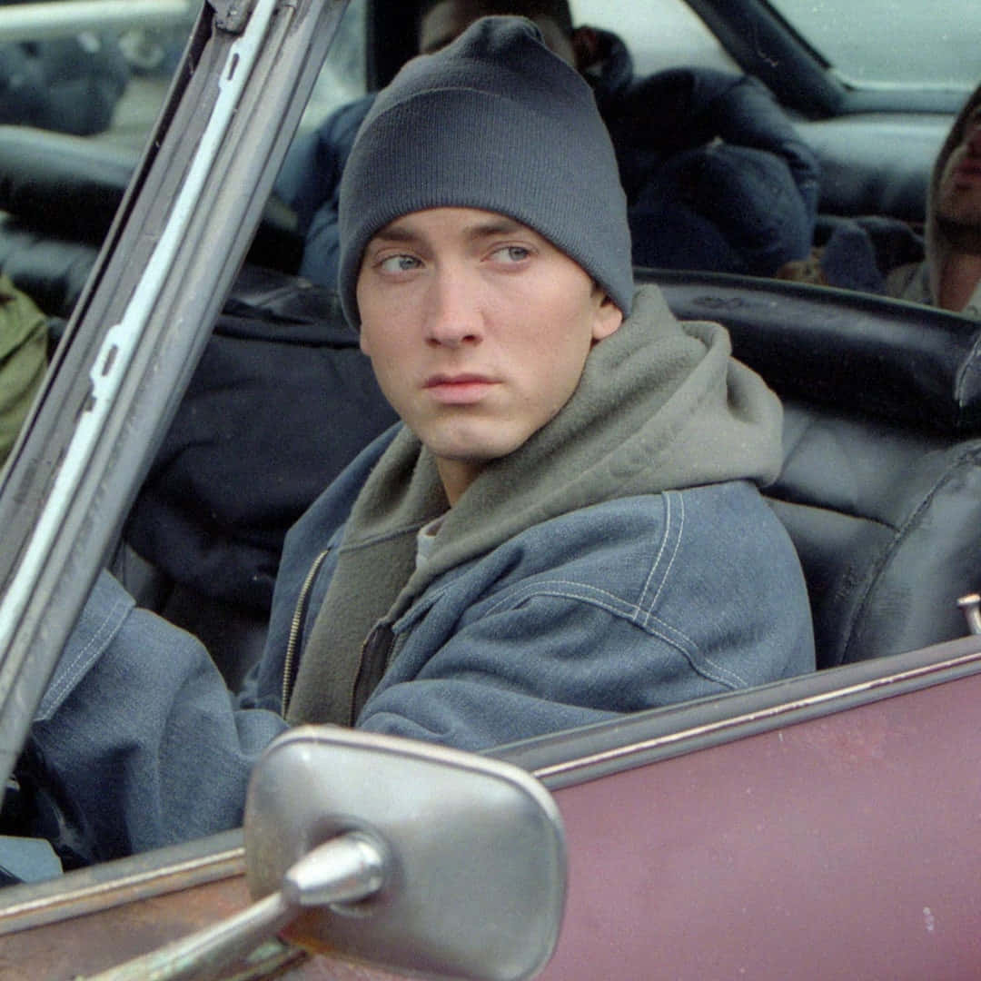 Eminem 桌布 壁紙 — 一個戴著冷帽和連帽衛衣的年輕男子，神情嚴肅地坐在一輛復古開篷車的駕駛座上。