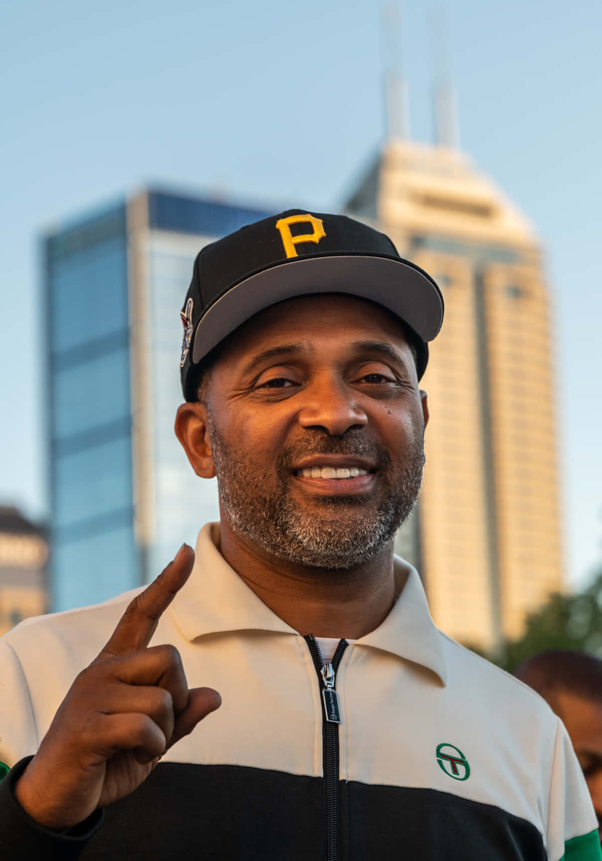 Mike Epps 桌布 壁紙 — 一個戴著匹茲堡海盜隊帽子的微笑男子，在晴朗藍天下以現代城市摩天大樓為背景向上指。