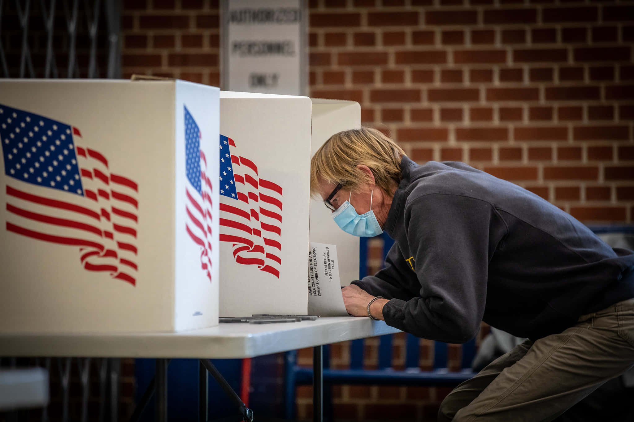 Election 桌布 壁紙 — 一個戴著口罩的人靠向一個裝飾著美國國旗圖案的投票亭，房間有磚牆。