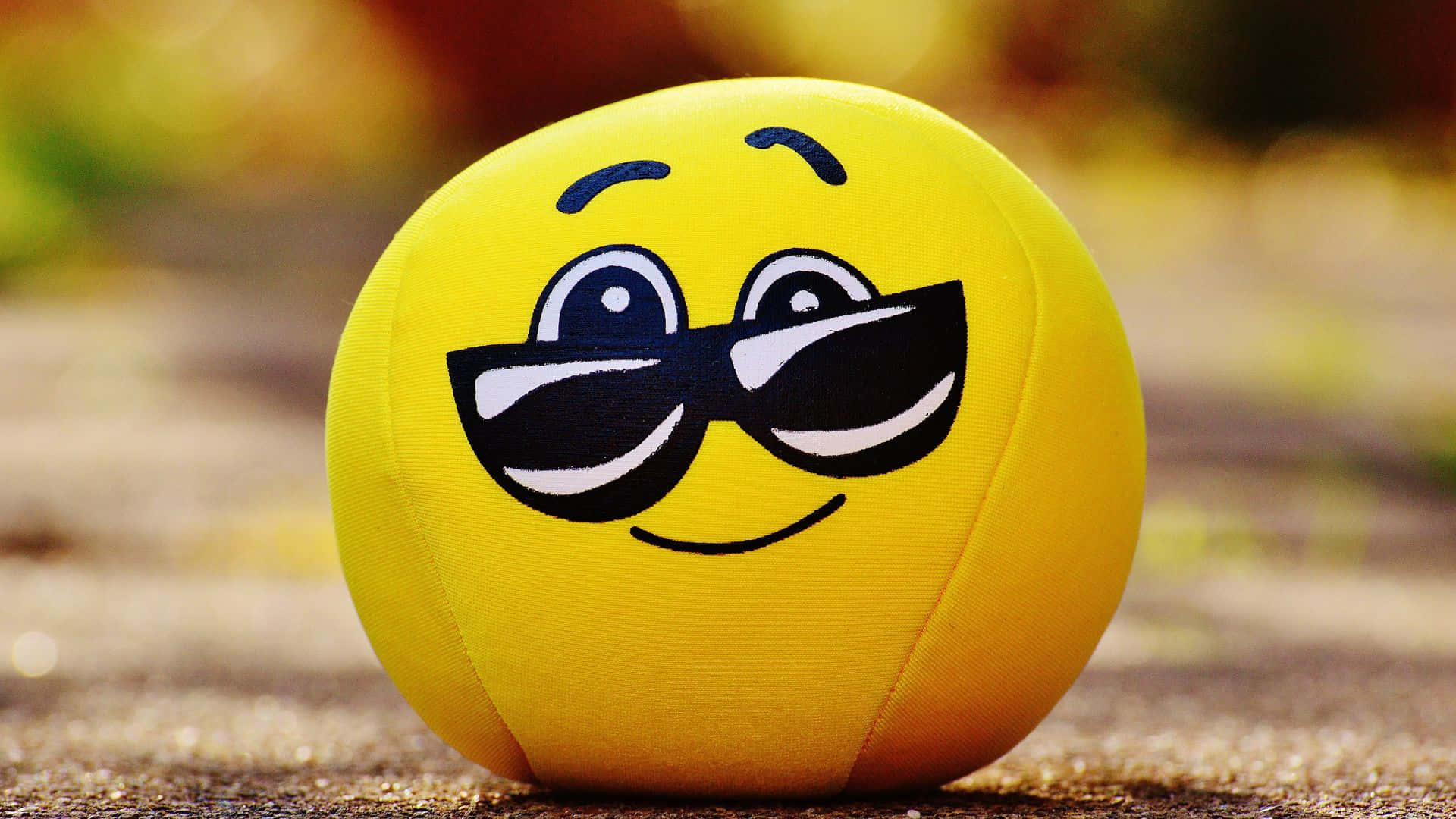 Smiley 桌布 壁紙 — 一個戴著型格太陽眼鏡的開心黃色表情符號波波，坐在沙地上，背後有模糊的秋葉。