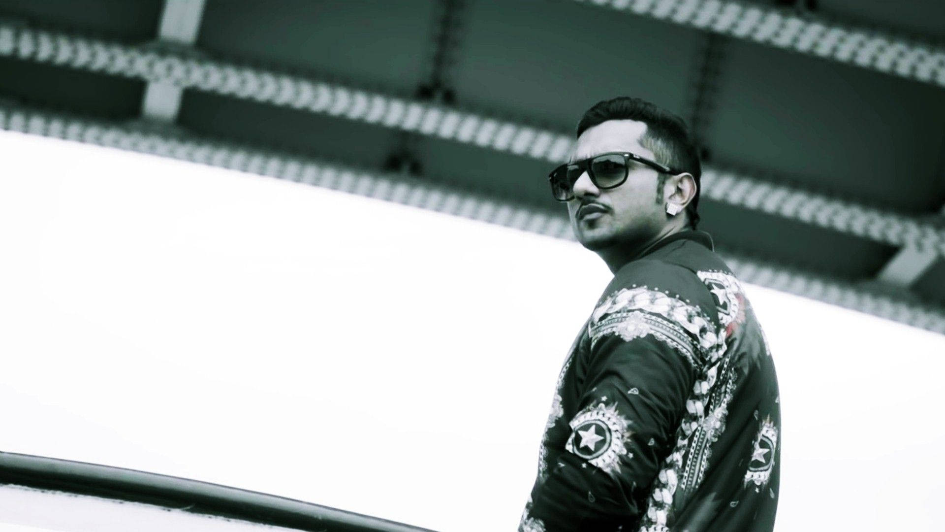 Yo Yo Honey Singh Call Aundi 壁紙 — 一個戴著太陽眼鏡和圖案外套的男人自信地站在一個線條強烈的簡約建築背景前。