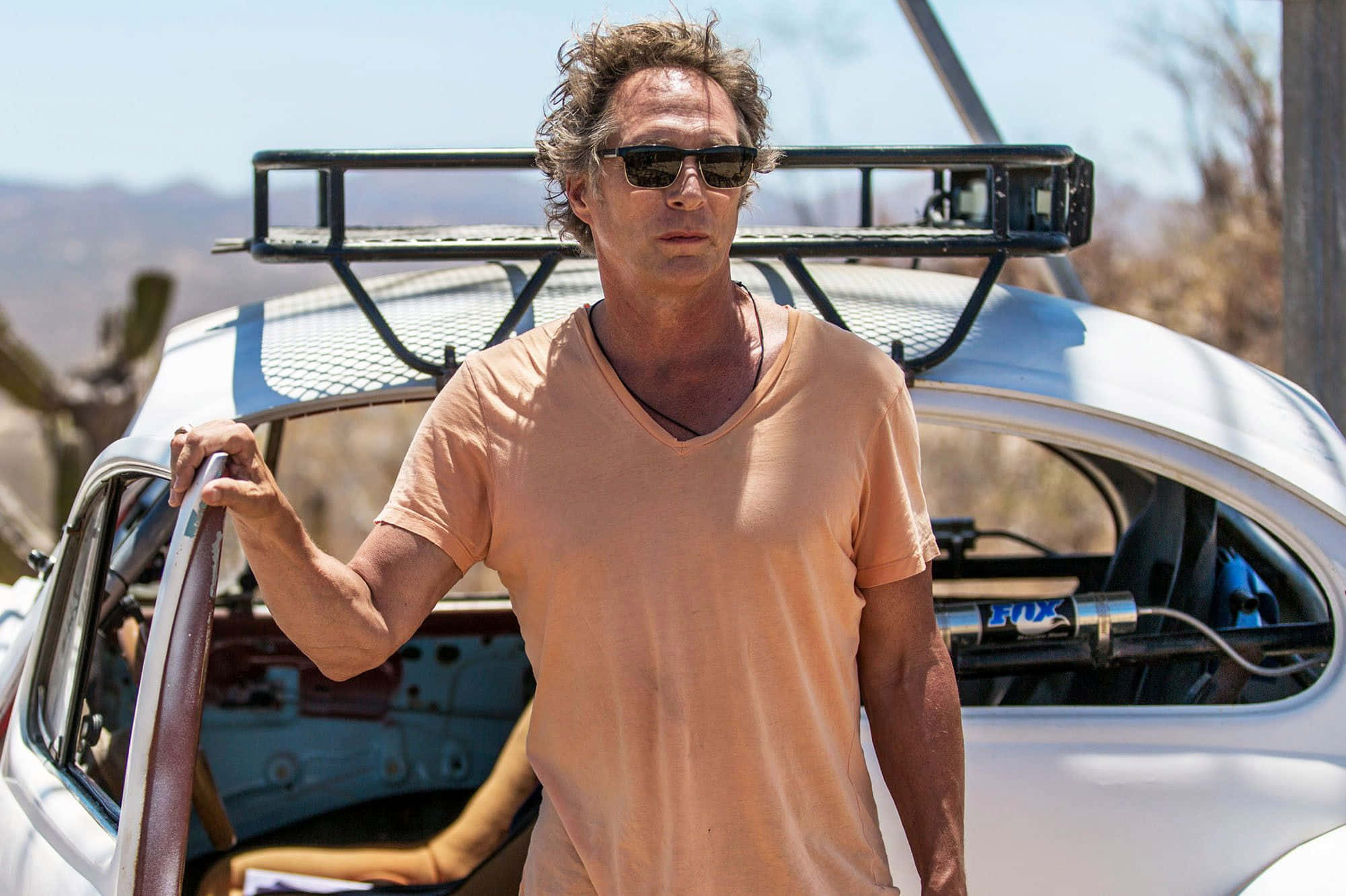 William Fichtner 桌布 壁紙 — 一個戴著太陽眼鏡的男人靠在一輛帶有車頂行李架的白色復古車上，背景是陽光普照的沙漠景觀。