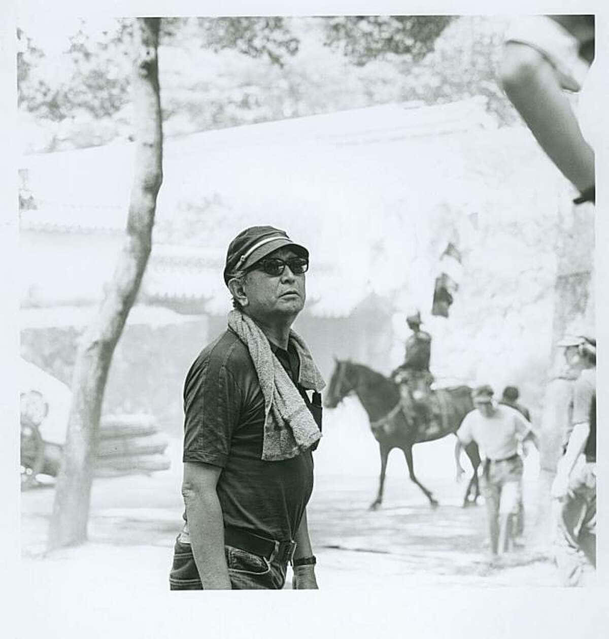 Akira Kurosawa 桌布 壁紙 — 一個戴著帽子和太陽眼鏡的男人站在陽光普照的戶外場景中，背景有馬匹和人。