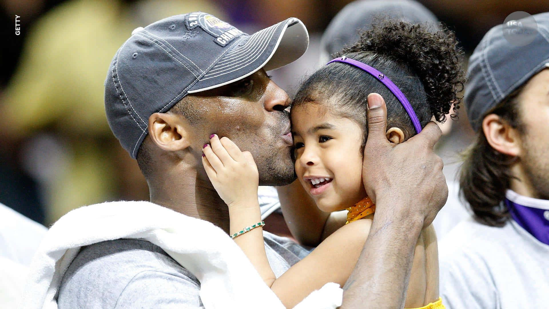 Kobe And Gigi 桌布 壁紙 — 一個戴著帽子的男人在體育賽事中抱著一個微笑的小女孩，親吻她的臉頰。
