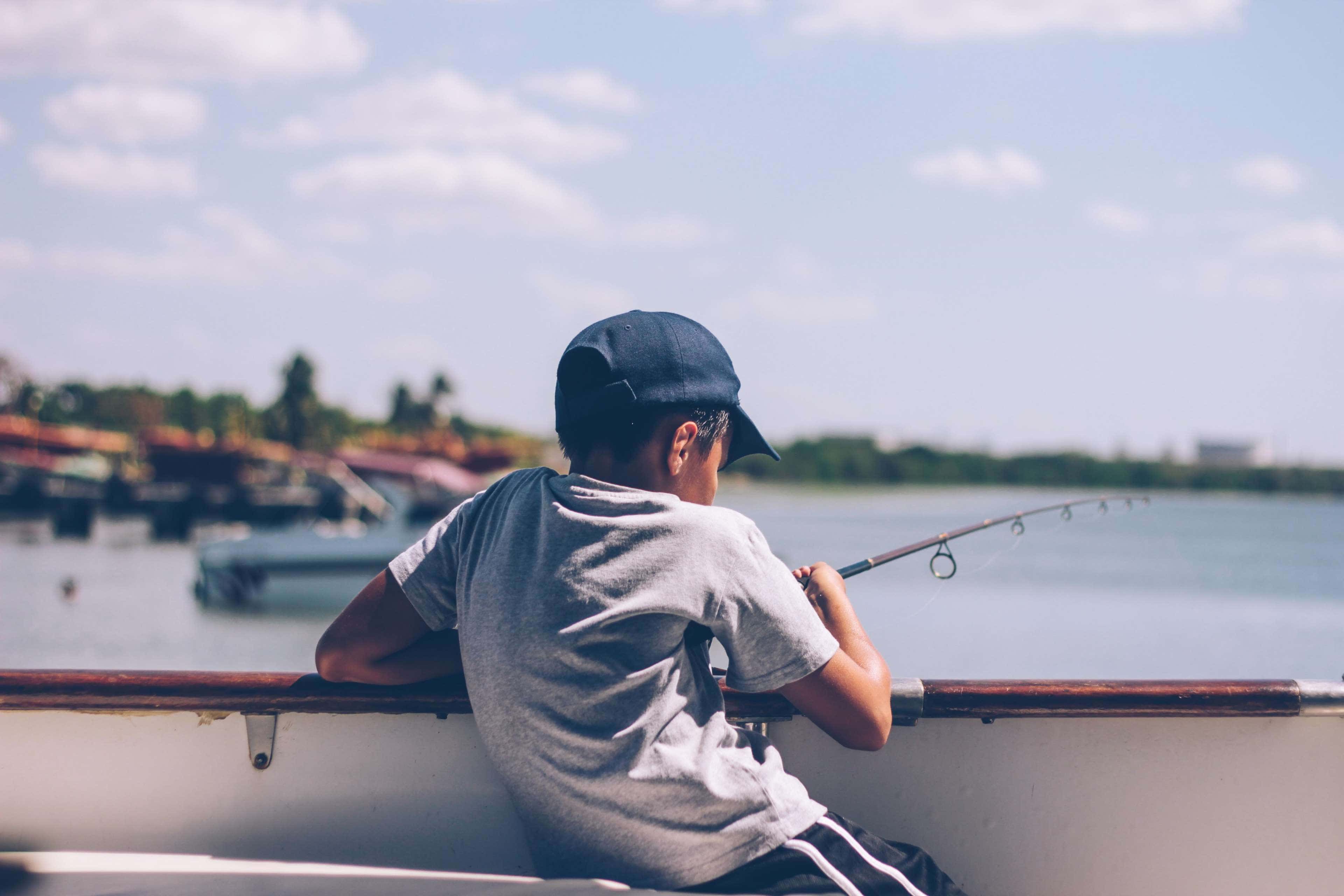 Fishing Bait 桌布 壁紙 — 一個戴著帽子的男孩坐在船上，拿著魚竿，凝視著部分多雲天空下的平靜水面。