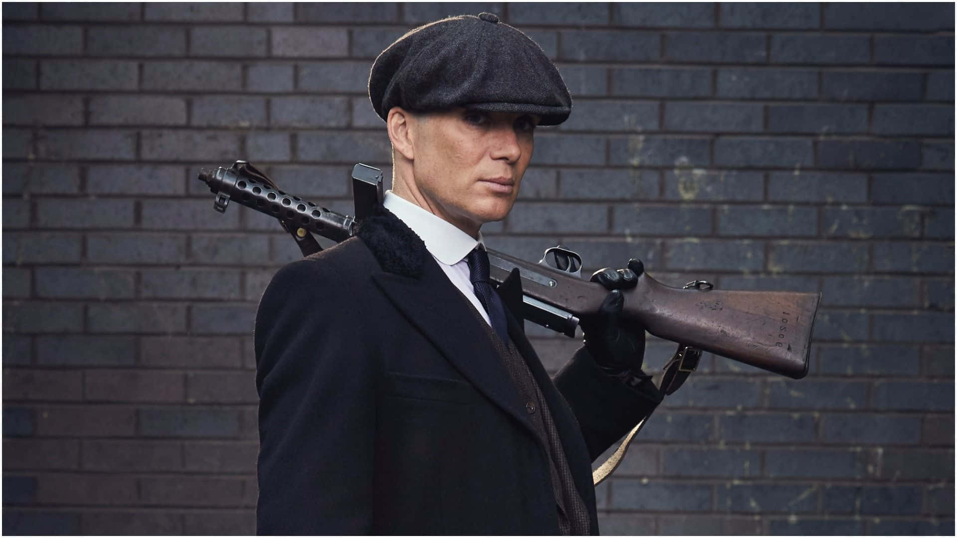 Peaky Blinders 桌布 壁紙 — 一個戴著平頂帽和風衣的男人，手持一把復古步槍，靠著深色磚牆，眼神凌厲。