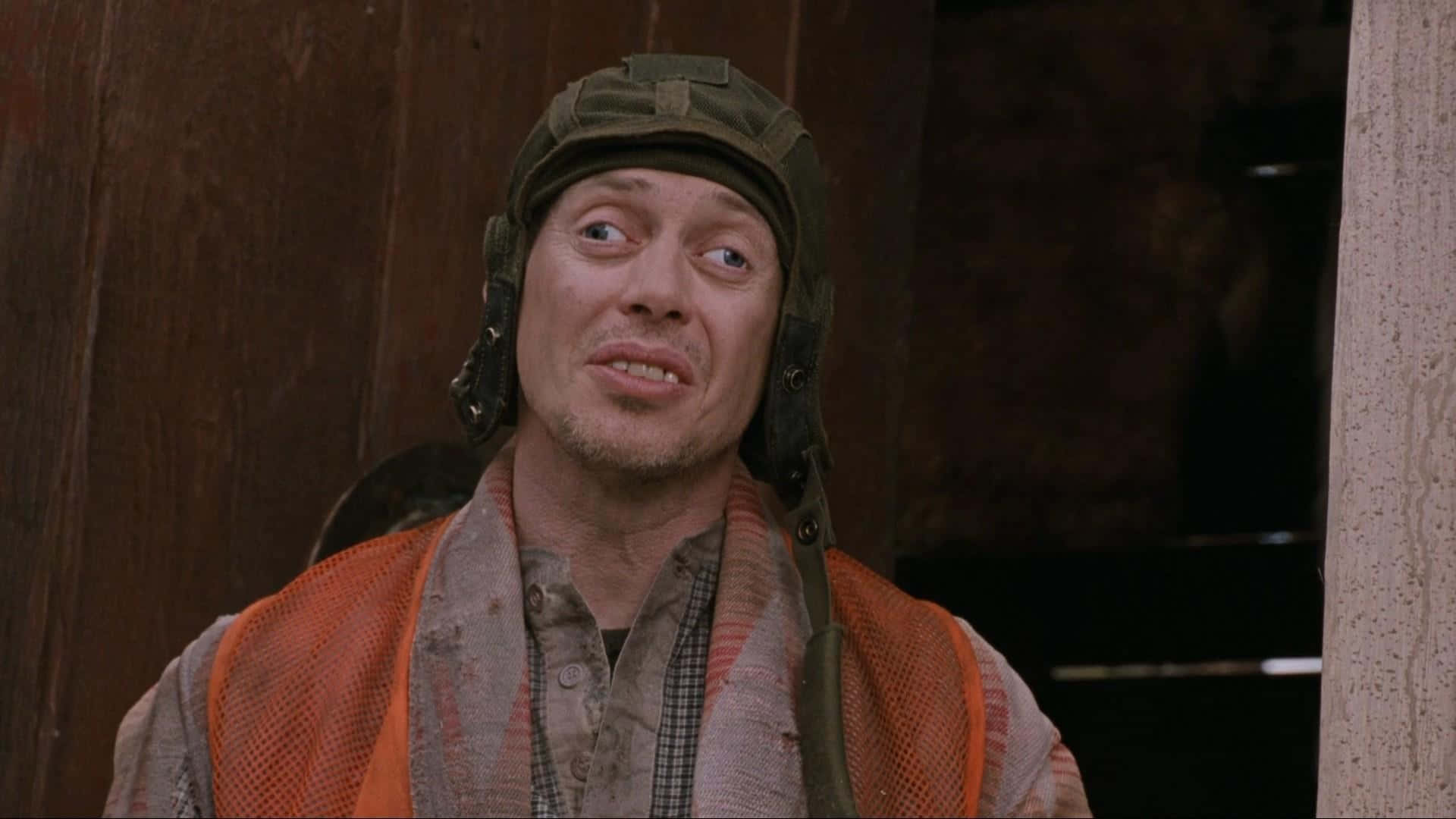 steve buscemi 桌布 壁紙 — 一個戴著泥濘飛行頭盔和橙色背心的男人，在木質背景下睜大眼睛，露出吃驚的表情。