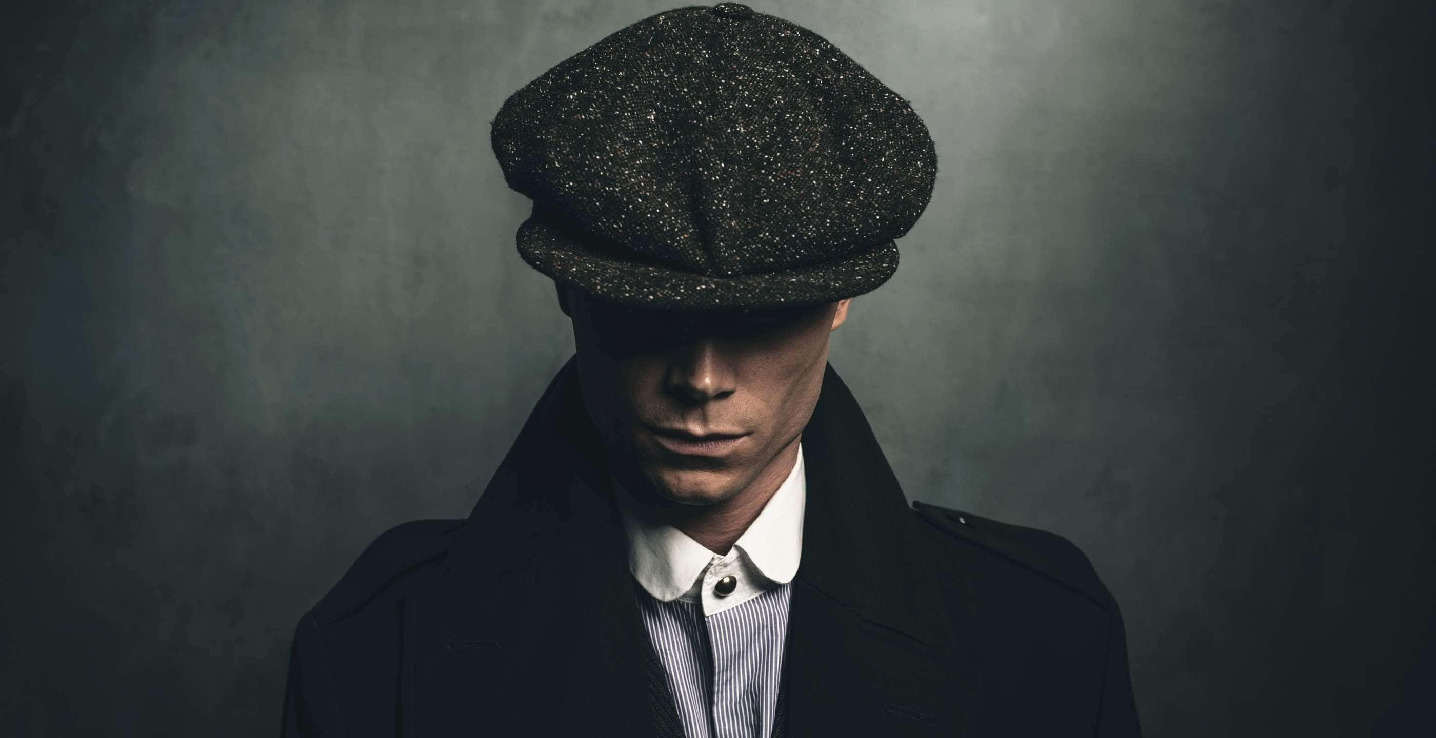 Peaky Blinders 桌布 壁紙 — 一個戴著深色平頂帽和外套的男人，在戲劇性燈光下，對著陰鬱的灰色背景專注地凝視。