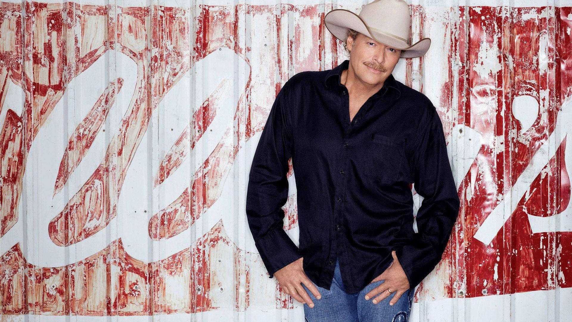 Alan Jackson 桌布 壁紙 — 一個戴著牛仔帽、身穿深色襯衫的男人靠在一面風化的紅白波紋金屬牆上，牆上有褪色的字樣。
