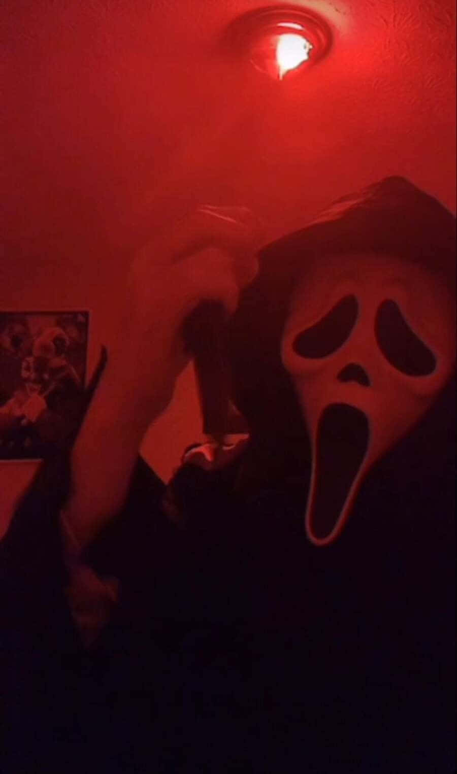 Ghostface Aesthetic 桌布 壁紙 — 一個戴著白色Ghostface面具的人，在昏暗房間的單一紅色天花燈下拿著一把刀。