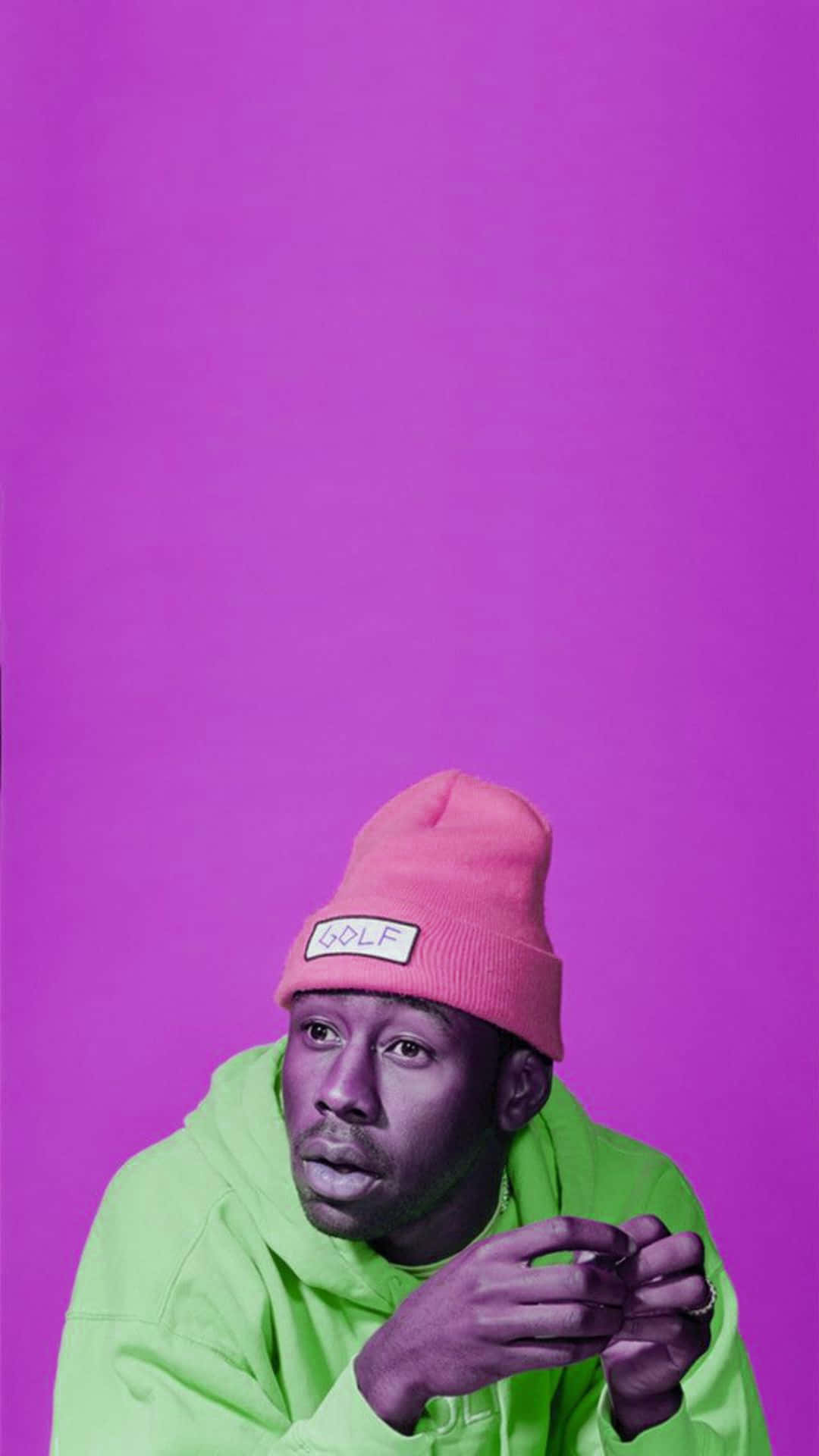 tyler the creator 桌布 壁紙 — 一個戴著粉紅色冷帽和螢光綠色連帽衫的男人，在純紫色背景前沉思地凝視著。