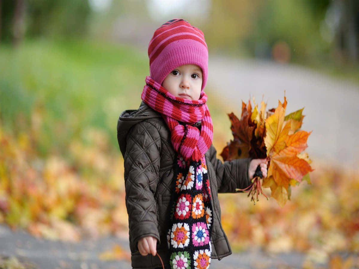 Beautiful Girls 桌布 壁紙 — 一個戴著粉紅色帽子和彩色圍巾的小孩，在鋪滿落葉的小路上拿著一束秋葉。