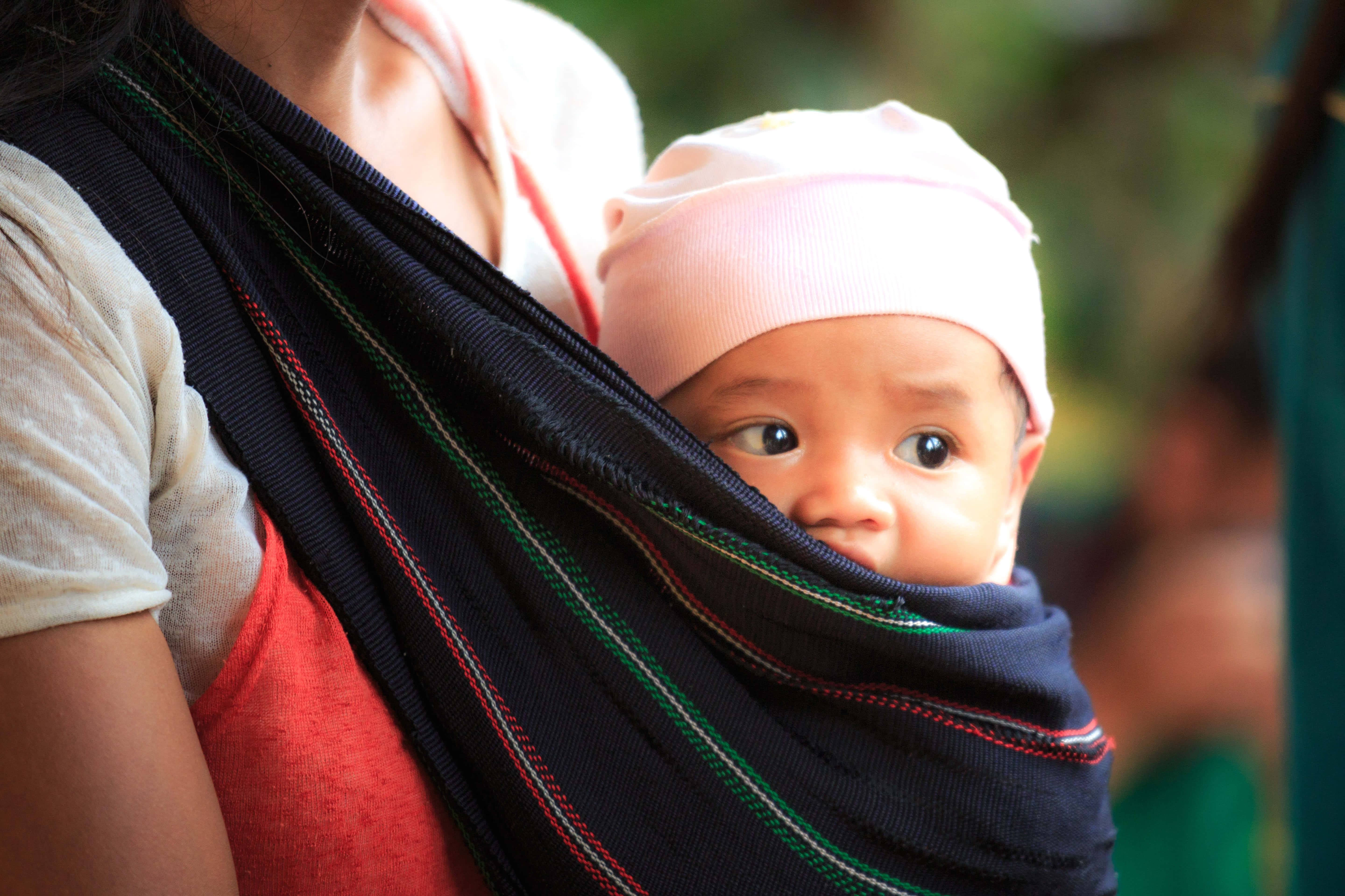 Mom 桌布 壁紙 — 一個戴著粉紅色帽子的嬰兒，從一個色彩繽紛的編織揹帶中探出頭來，由一位成年人在茂密的綠色戶外環境中背著。