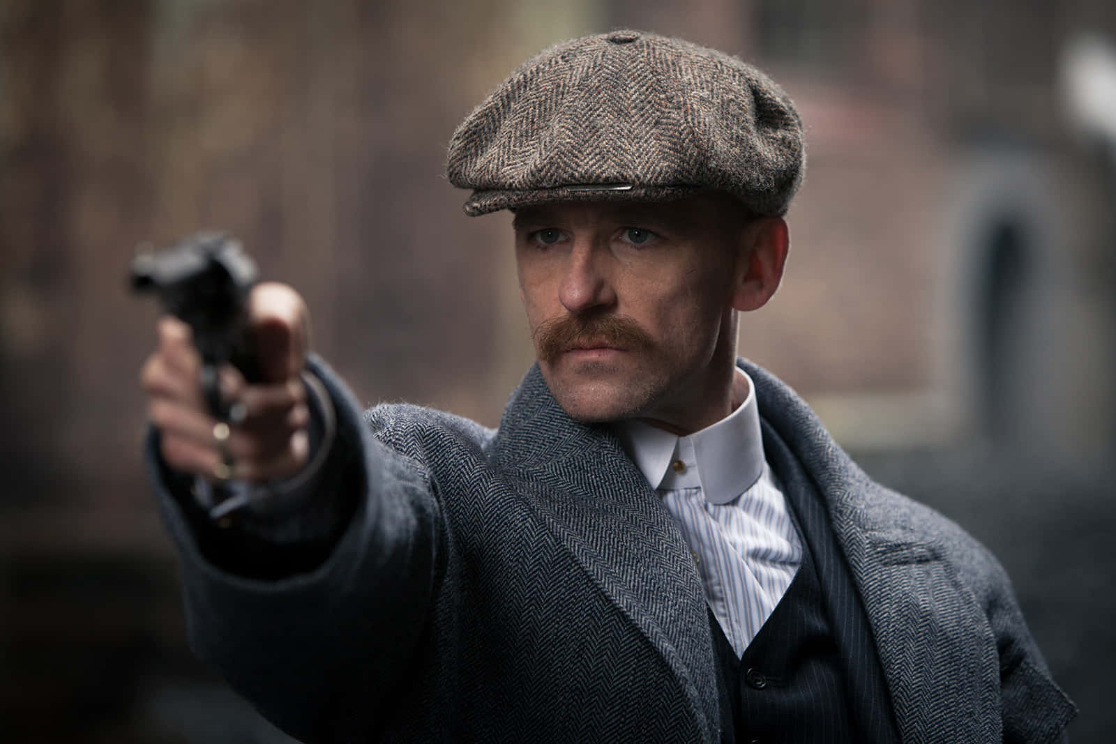 Peaky Blinders 桌布 壁紙 — 一個戴著粗花呢平頂帽和穿著外套的男人，在模糊的磚牆背景下，以嚴厲的目光瞄準一把手槍。
