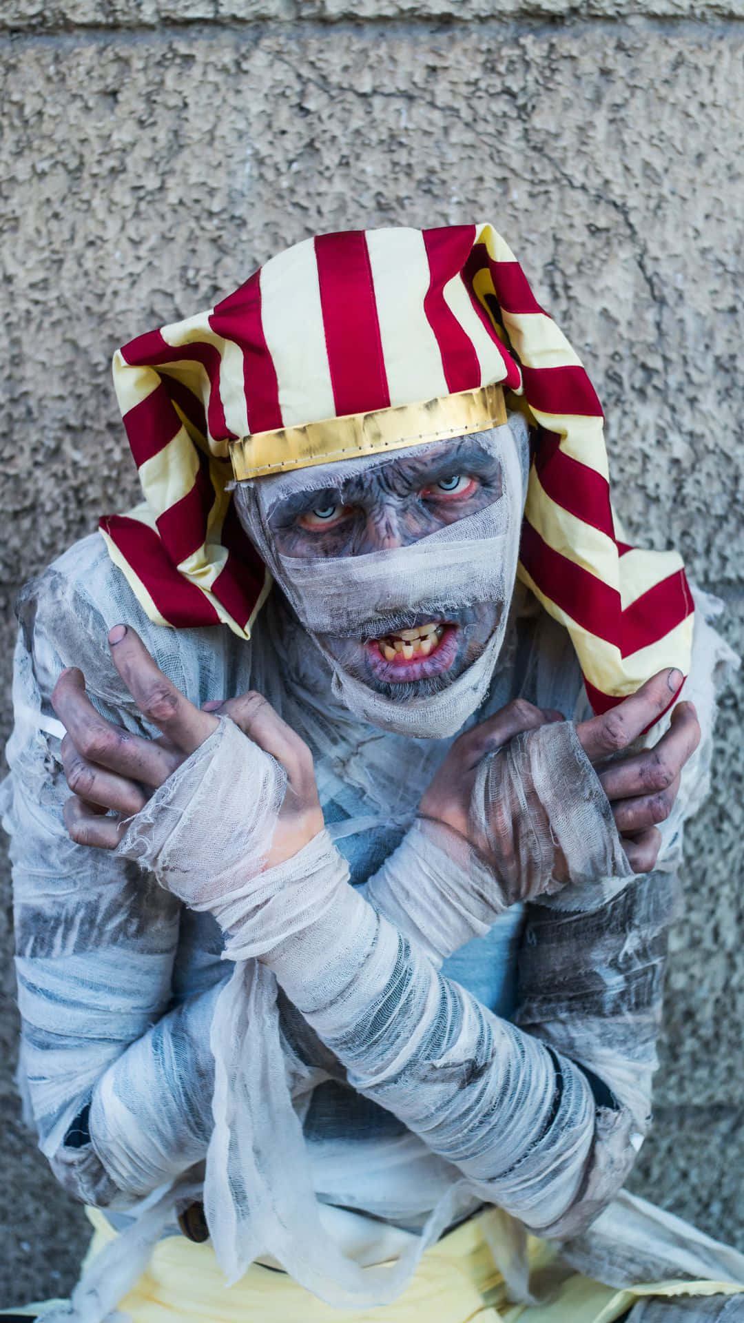 Mummy Costumes 桌布 壁紙 — 一個戴著紅白條紋帽子、臉部纏著繃帶的怪誕木乃伊，正對著粗糙的混凝土牆壁凝視。