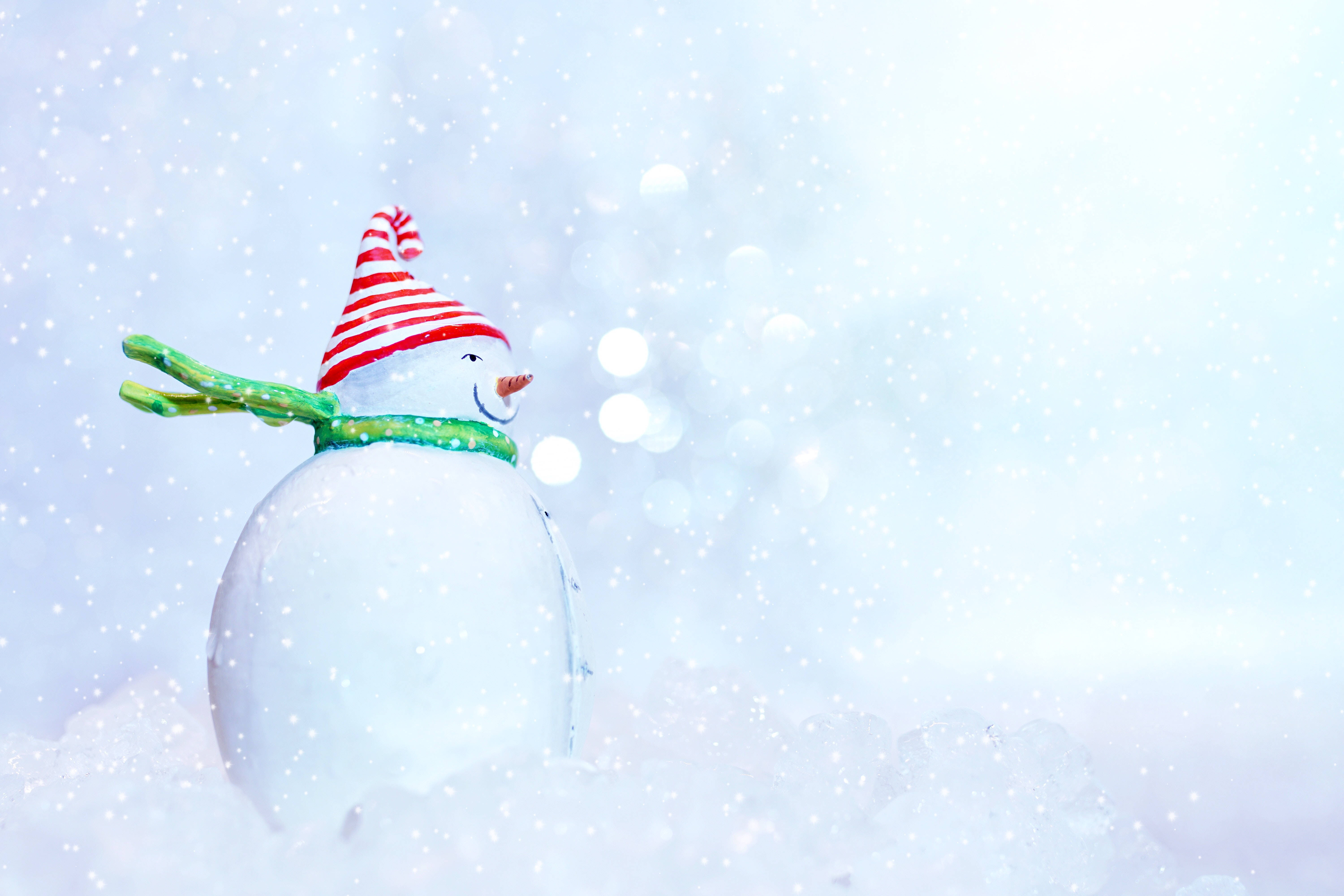 雪人 桌布 壁紙 — 一個戴著紅白條紋帽子和綠色圍巾的快樂雪人，站在飄雪和柔和散景燈光的雪景中。