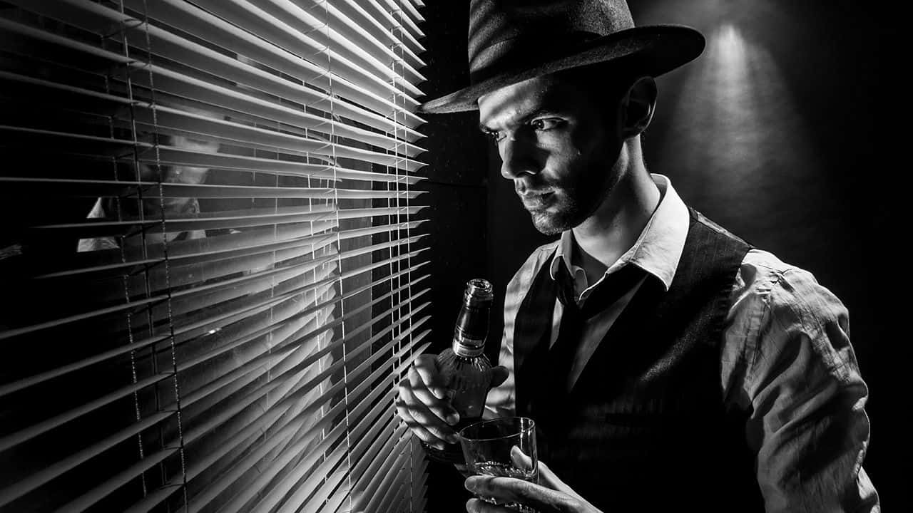 Film Noir 桌布 壁紙 — 一個戴著紳士帽的男人，在單色調的戲劇性聚光燈下，拿著一個瓶子和一個玻璃杯，旁邊是百葉窗。