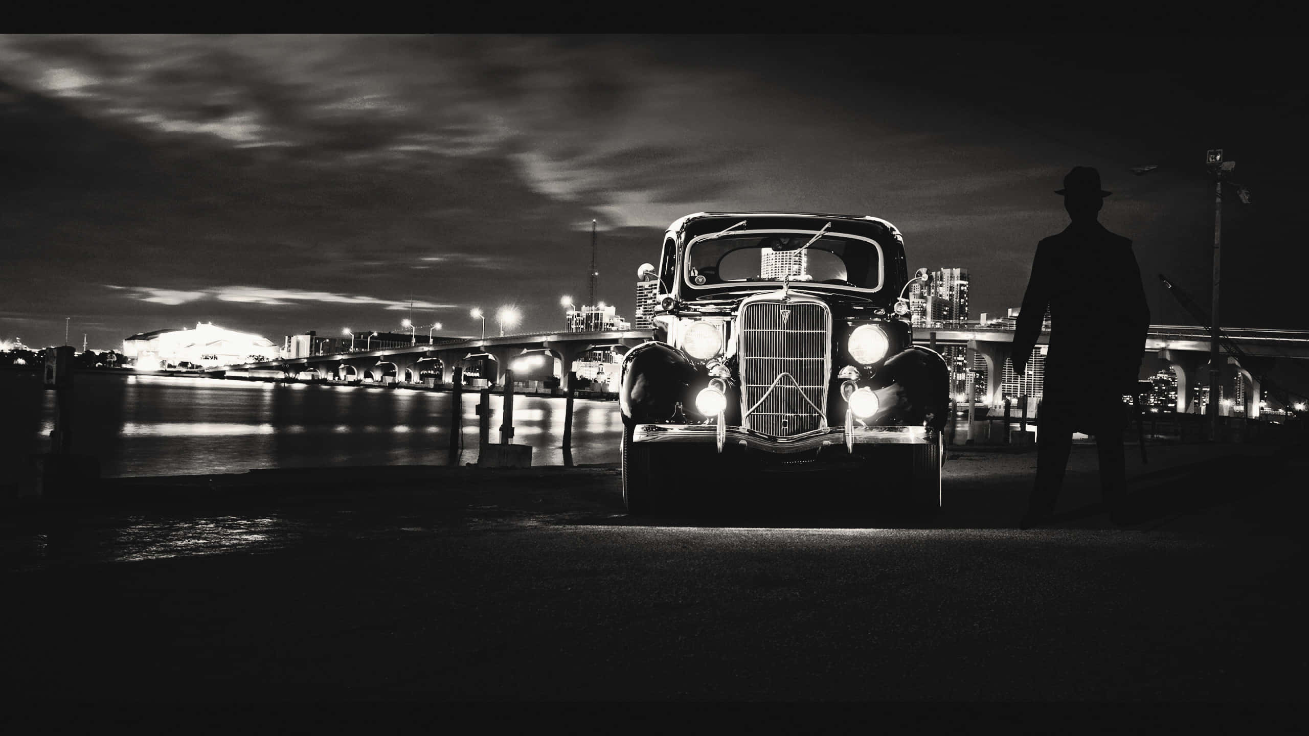 Black And White Movie 桌布 壁紙 — 一個戴著紳士帽的男子的黑色剪影，站在一輛車頭燈發光的復古汽車旁，背景是夜晚的海濱。