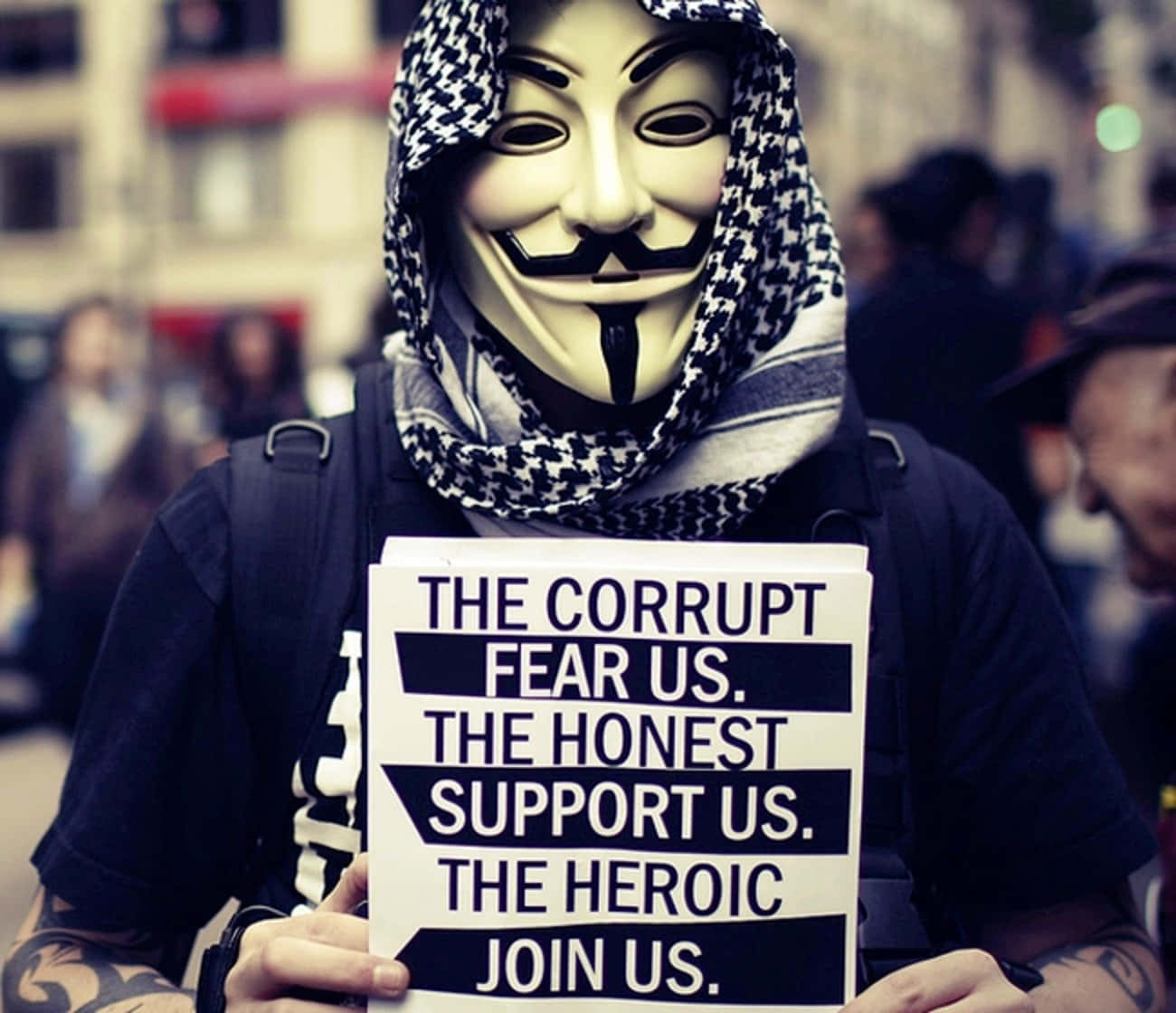 Anonymous 桌布 壁紙 — 一個戴著蓋伊·福克斯面具和圖案圍巾的人，在擁擠的城市街道上舉著抗議標語。