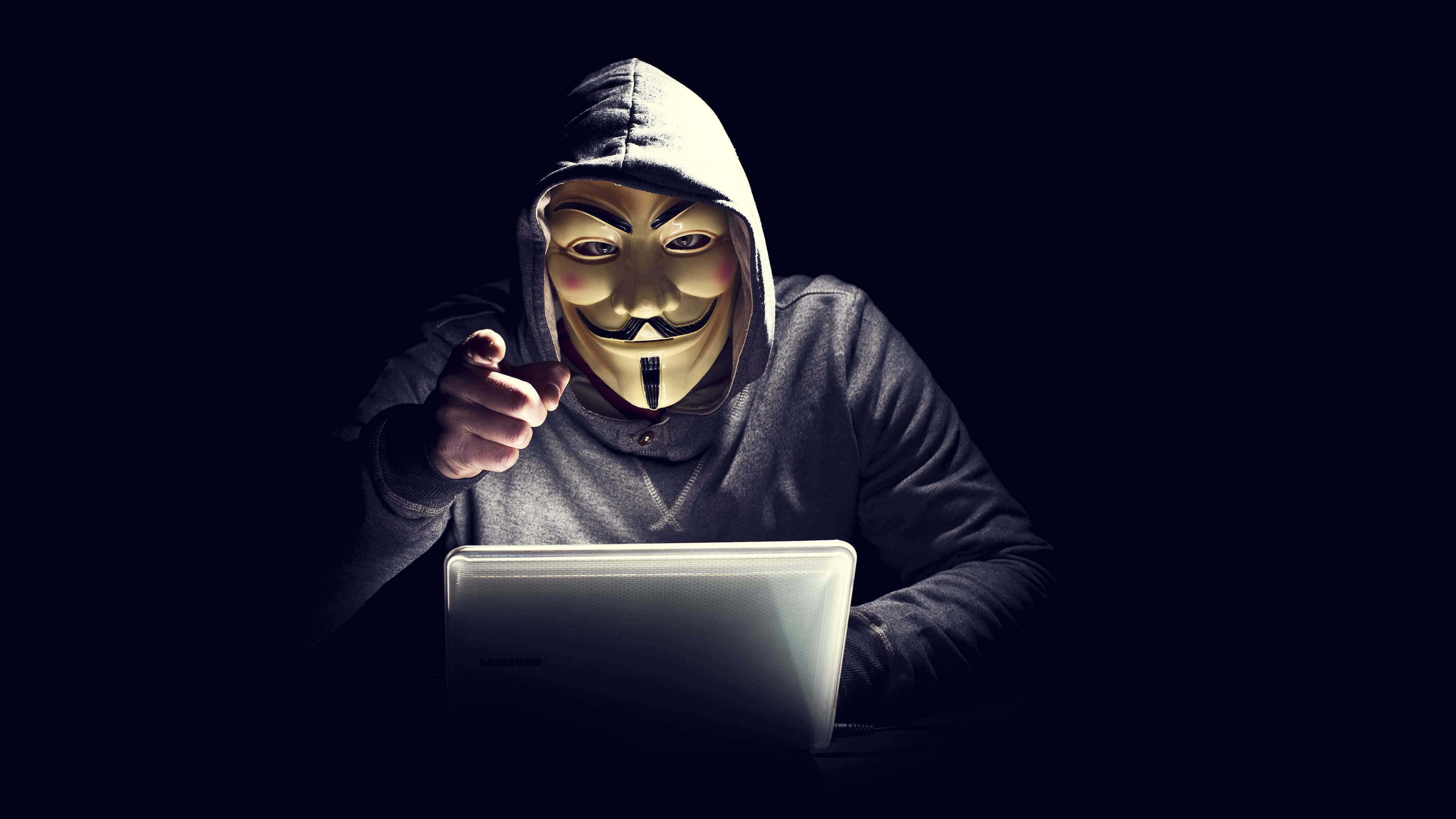 Anonymous 桌布 壁紙 — 一個戴著蓋伊·福克斯面具和連帽衫的人，在黑暗的房間裡，被屏幕的光芒照亮，指責地指著一部筆記本電腦。