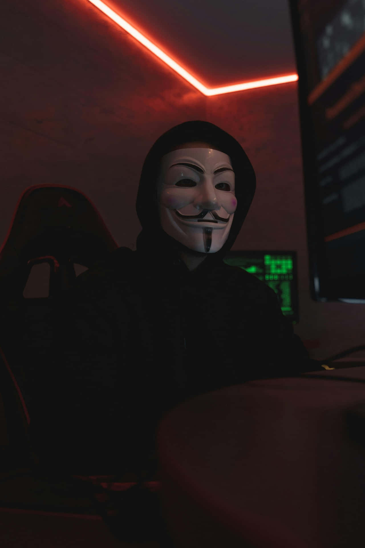  壁紙 — 一個戴著蓋伊·福克斯面具的人坐在一個昏暗的房間裡，房間被天花板的紅色霓虹燈條和發光的電腦屏幕淹沒。