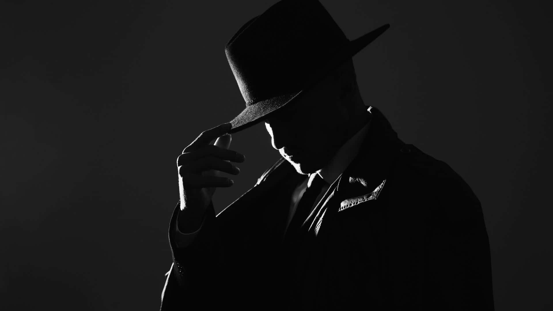 Black And White Movie 桌布 壁紙 — 一個戴著軟呢帽和風衣的影子般的男人在戲劇性的背光下調整他的帽子。