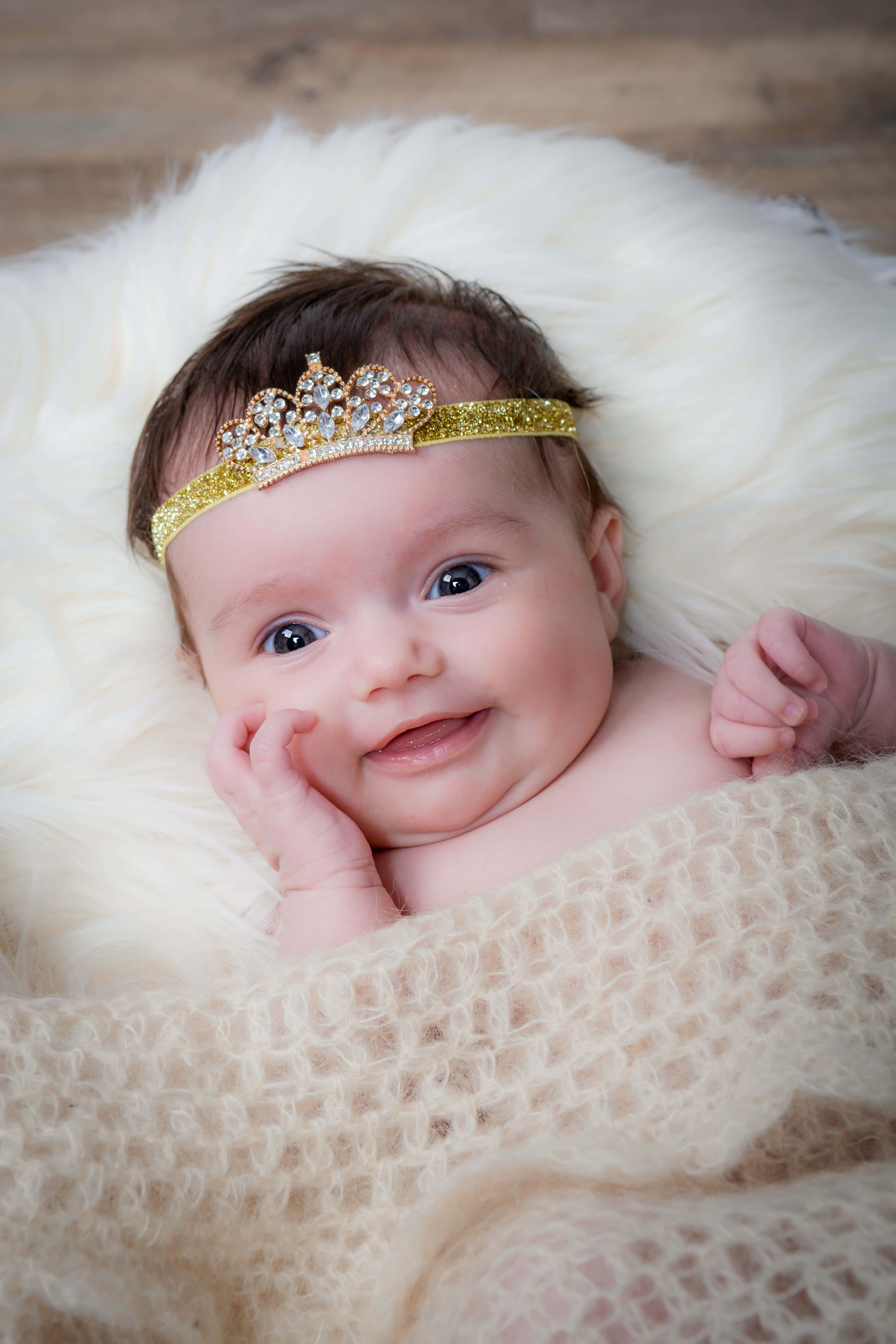 Baby Girl 桌布 壁紙 — 一個戴著金色頭飾的微笑嬰兒在柔和燈光下躺在柔軟的白色地毯上休息。