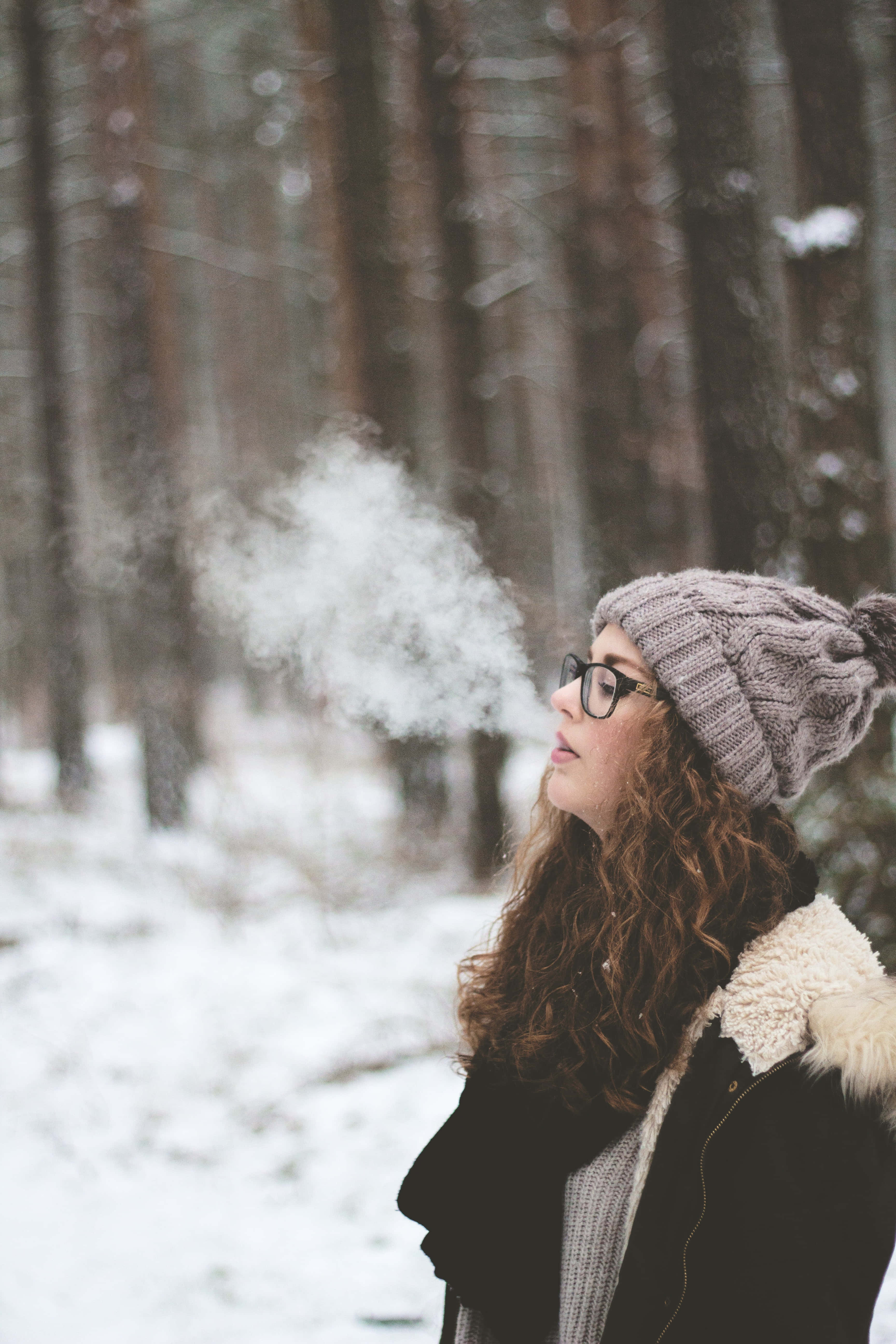 寒冷 桌布 壁紙 — 一個戴著針織帽的女人向寒冷積雪的森林呼出溫暖的氣息，她的眼鏡起霧，捲髮勾勒出她的臉龐。