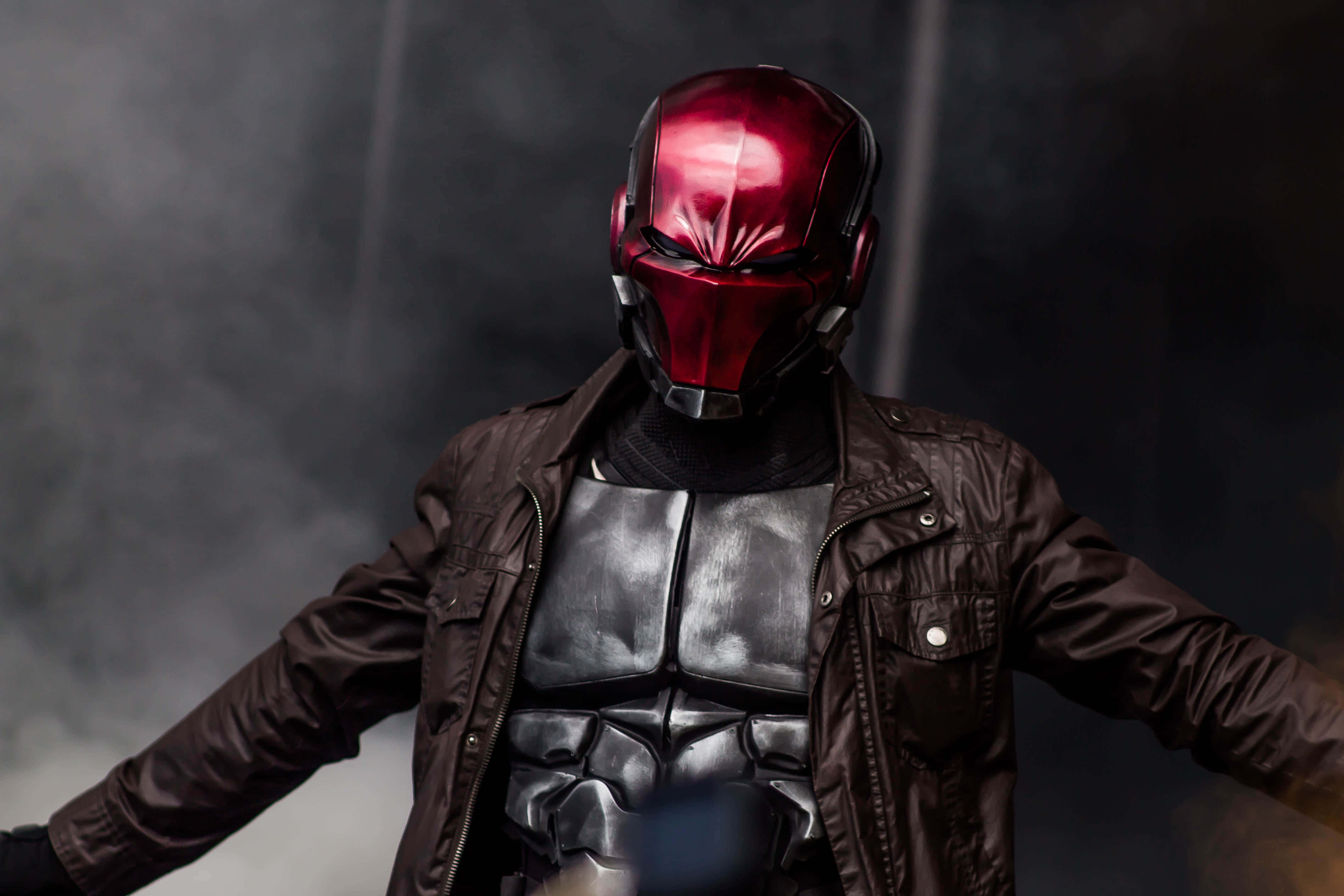 Red Hood 桌布 壁紙 — 一個戴著閃亮紅色頭盔、身穿黑色裝甲服的人物，在煙霧瀰漫的黑暗背景下張開雙臂站立。