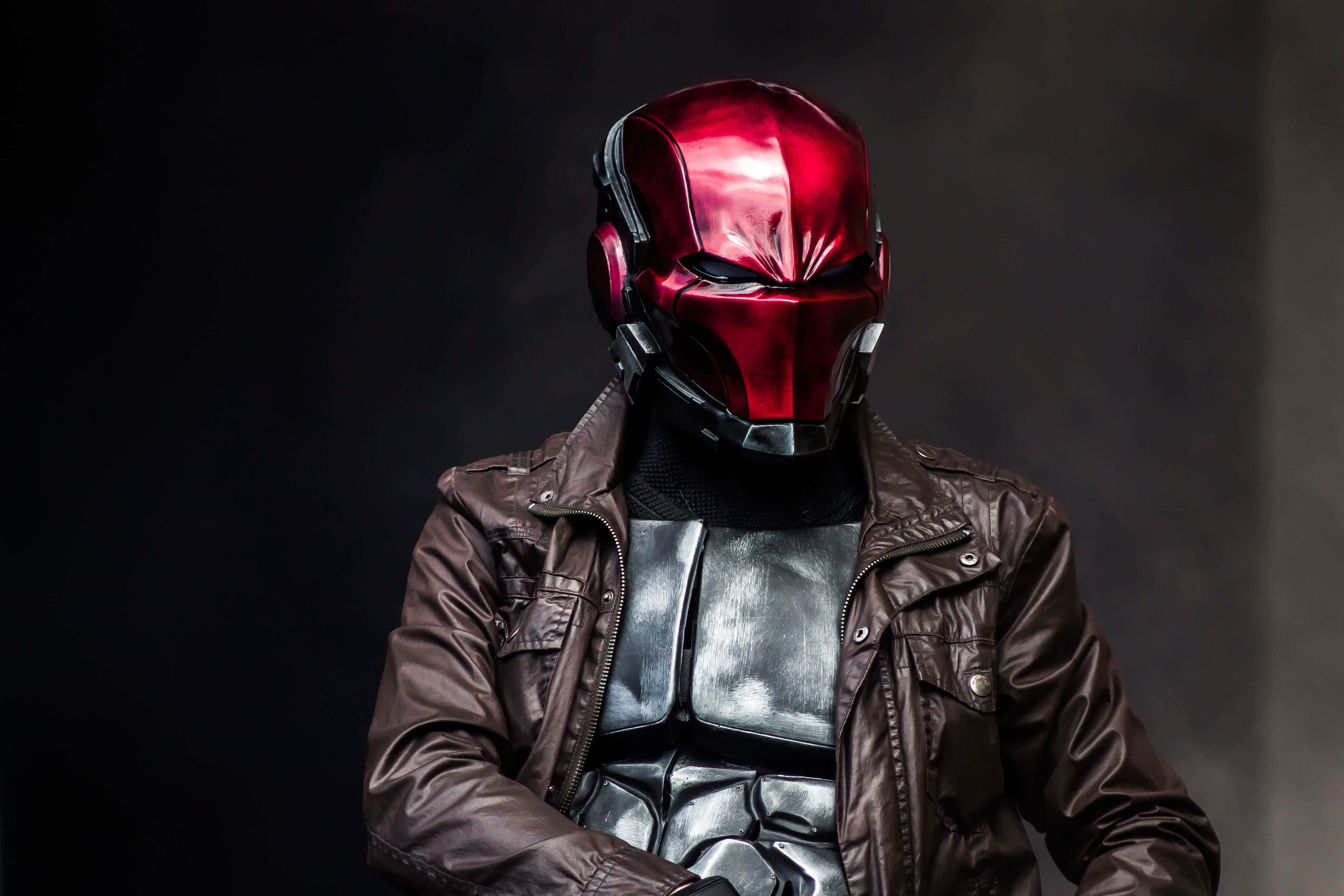 Red Hood 桌布 壁紙 — 一個戴著閃亮紅色頭盔和棕色皮褸的人物站在黑暗煙霧背景前。