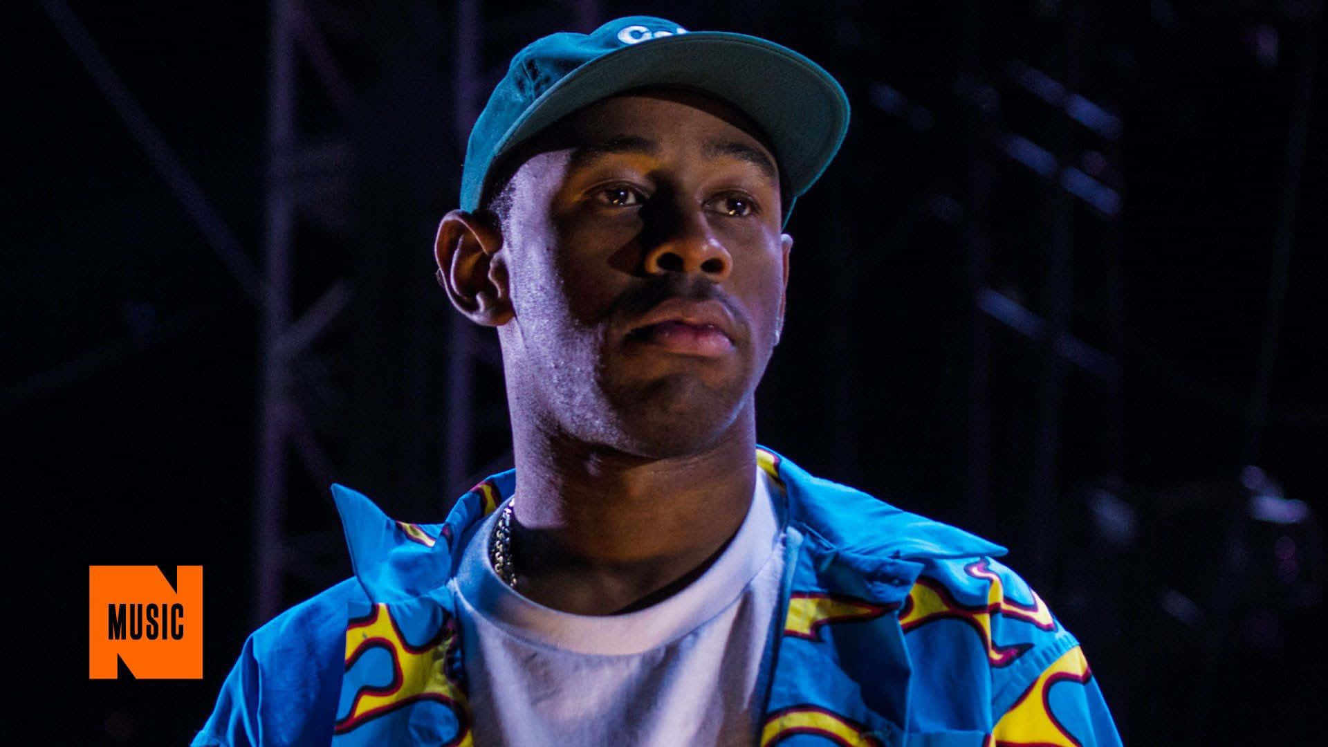 tyler the creator 桌布 壁紙 — 一個戴著青綠色帽子和鮮藍色外套的男人在舞台燈光下仰望黑暗背景。